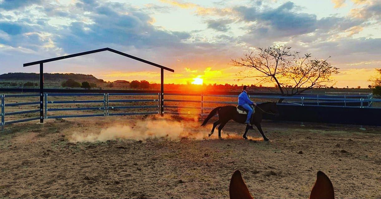 A man is riding a horse in a fenced in area at sunset.