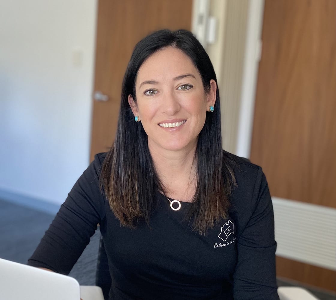 A woman is sitting at a desk with a laptop and smiling.