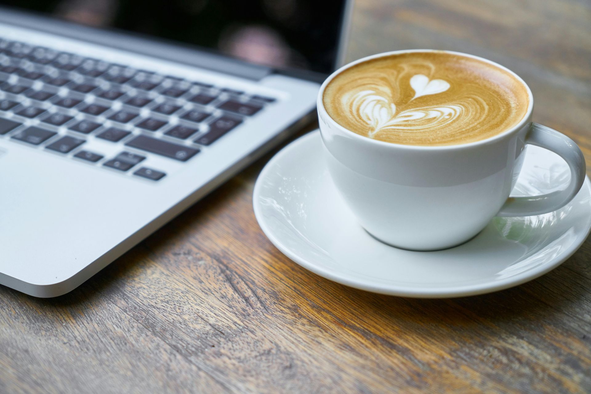 A cup of coffee on a saucer next to a laptop on a wooden table.