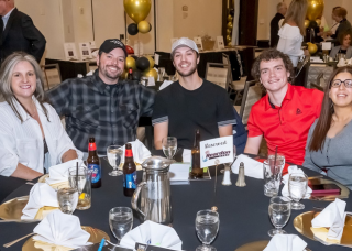 A group of six people smiling at a table, seated at an event. They are surrounded by decorations and food.