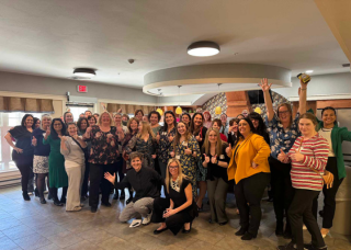 Group of five people smiling at a table, possibly at a banquet. They are surrounded by glasses, beer bottles, and gold accents.