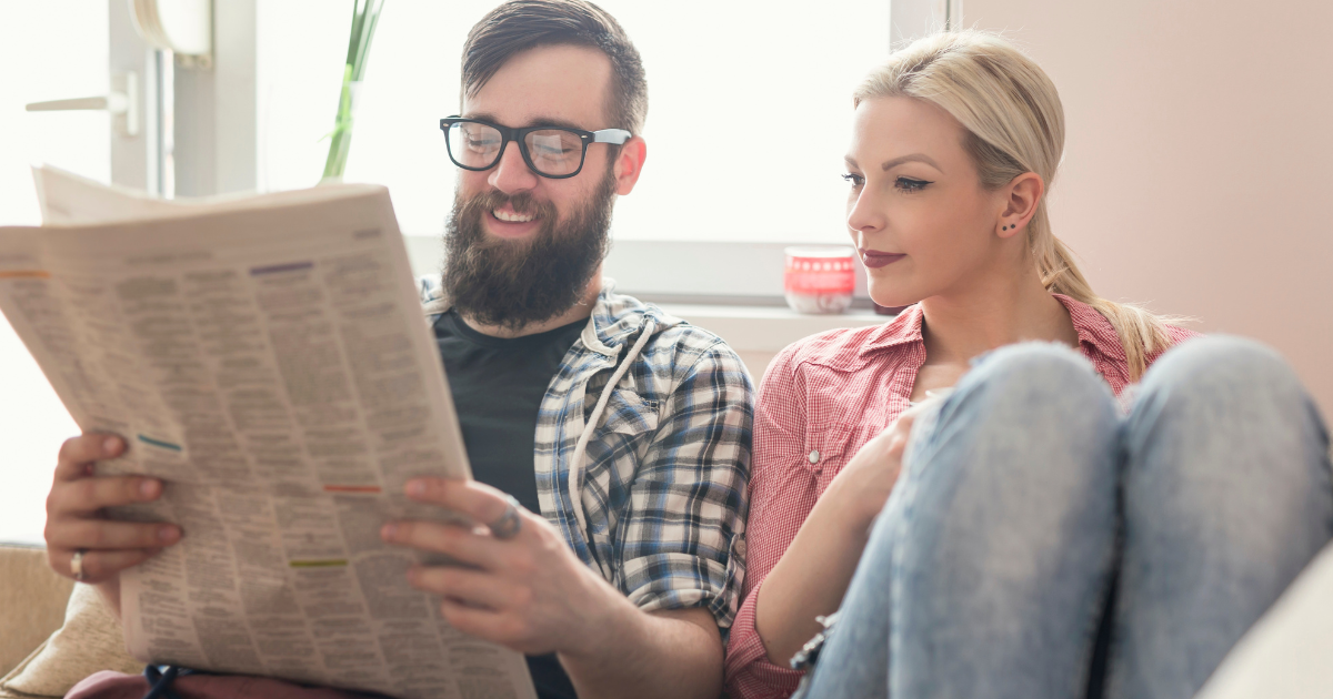 A man and a woman are sitting on a couch reading a newspaper.