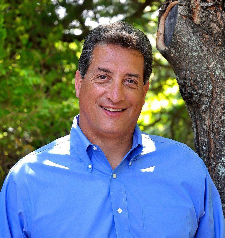 A man in a blue shirt is smiling in front of a tree