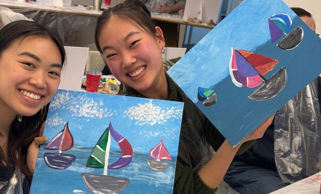 Two smiling young women hold up their colorful sailboat paintings, showing off their art in a bright room.