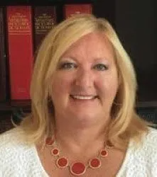 A woman wearing a white shirt and a red necklace is smiling in front of a bookshelf.