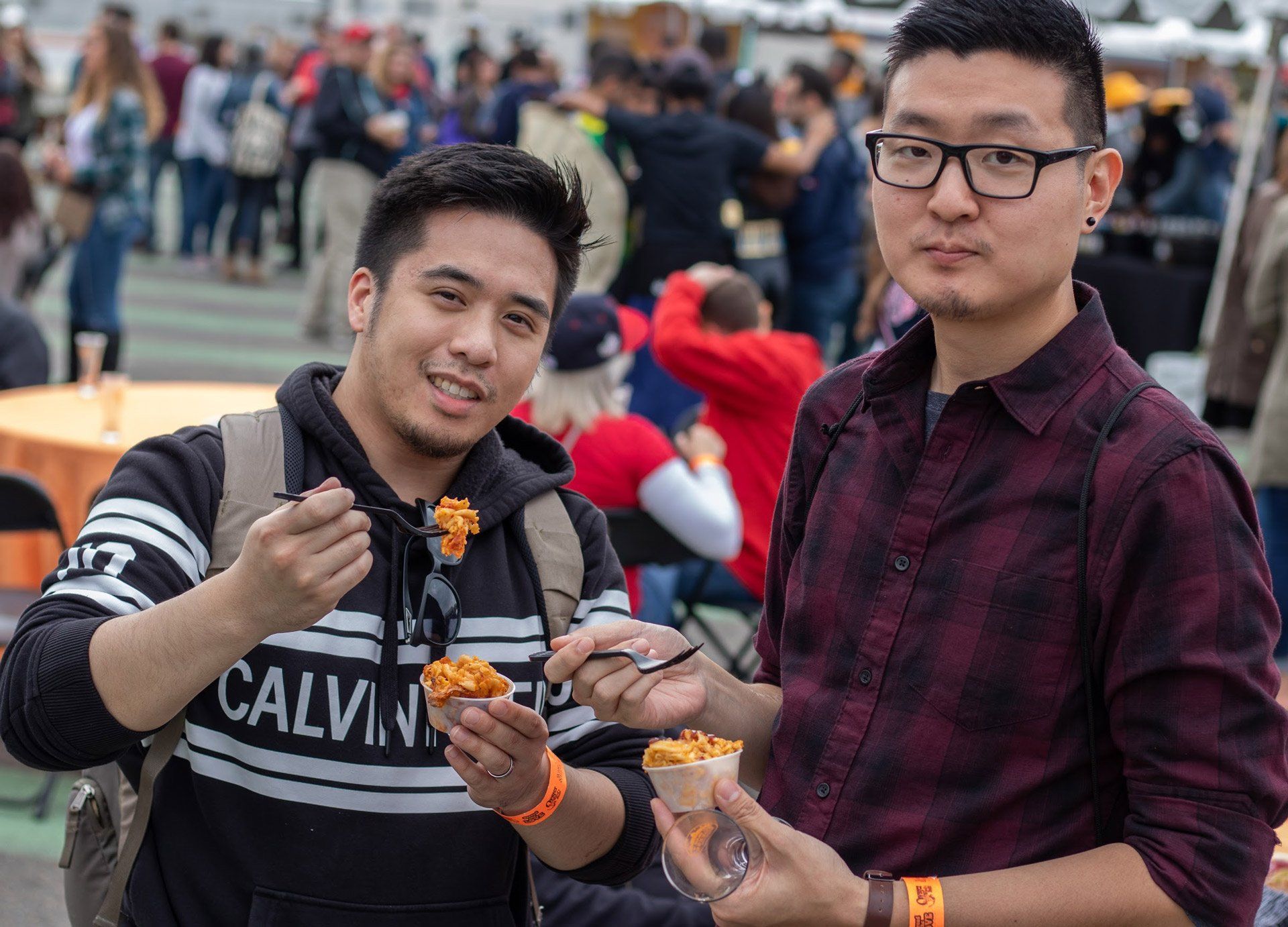 Two men are standing next to each other eating food at a festival.