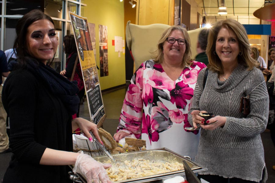 Three women are standing around a tray of food.