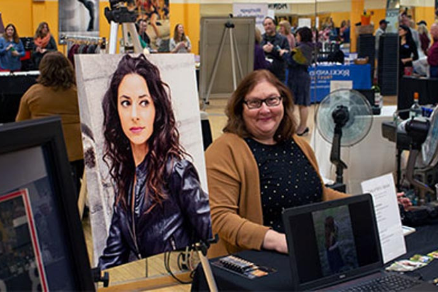 A woman is sitting at a table with a laptop and a painting of a woman.