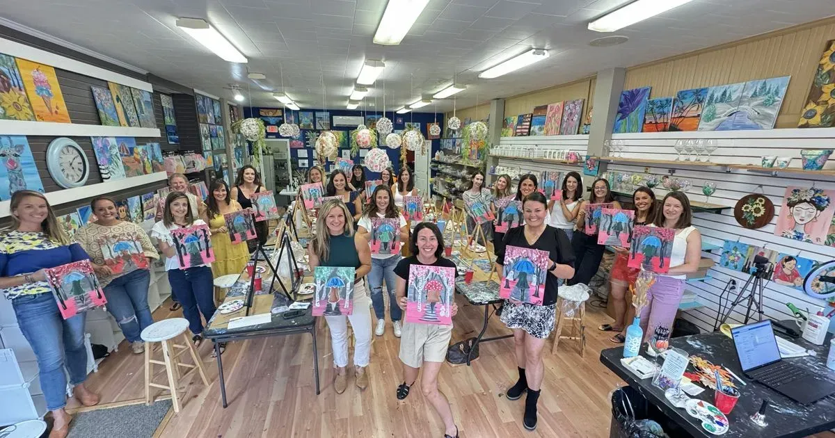 Group of people in an art studio, holding up paintings with smiling expressions; paintings displayed on walls.