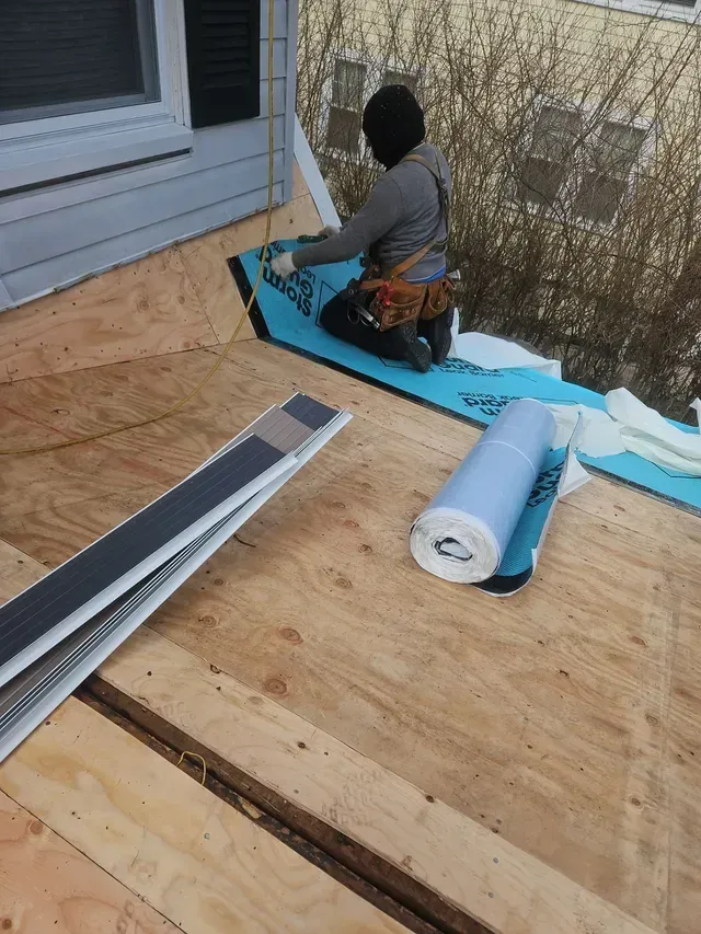 Roofer installing a blue underlayment on a plywood roof, with rolls of material and roofing supplies visible.