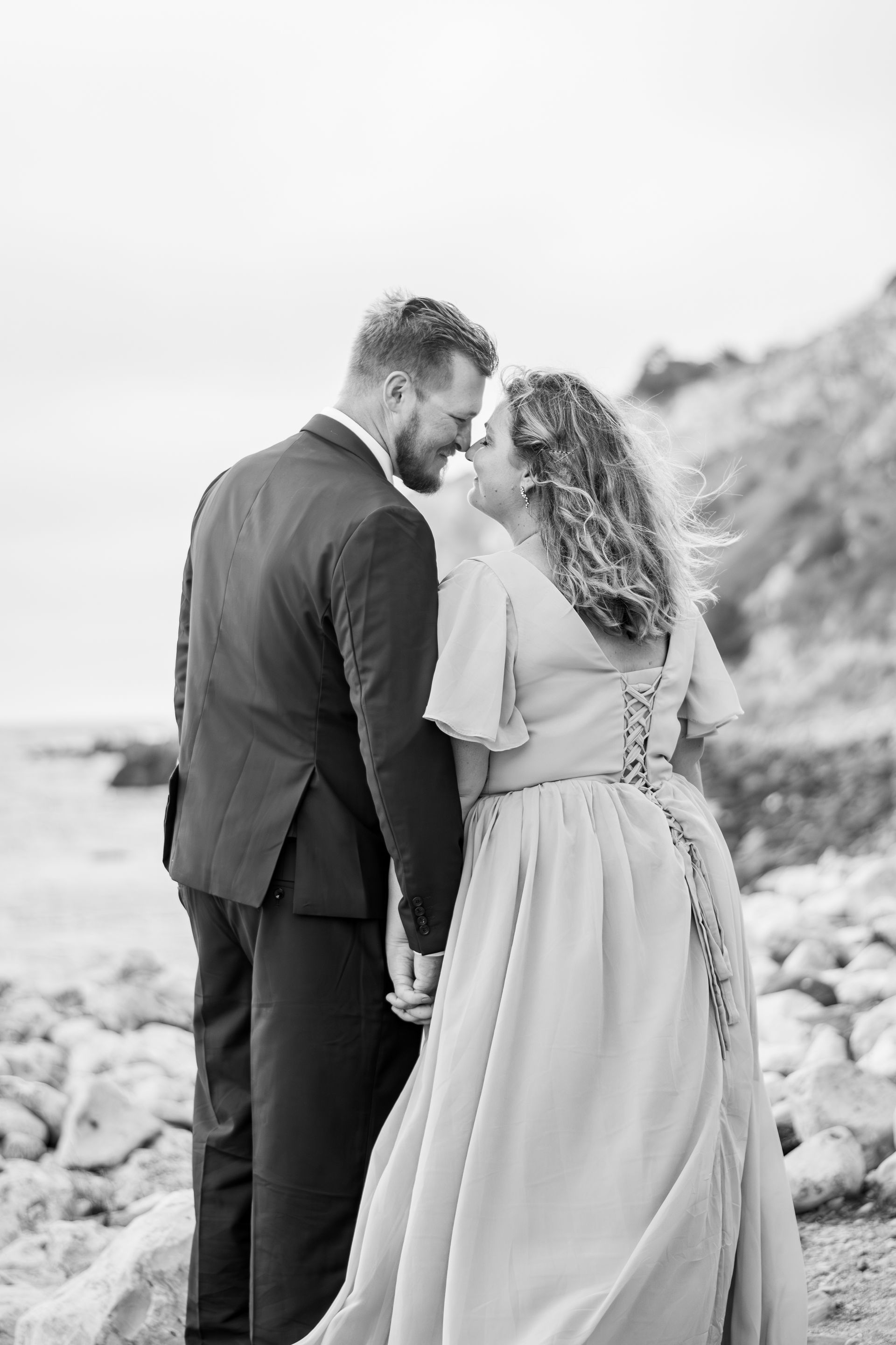 A couple in formal attire holds hands while facing each other on a rocky beach by the ocean. Black and white.