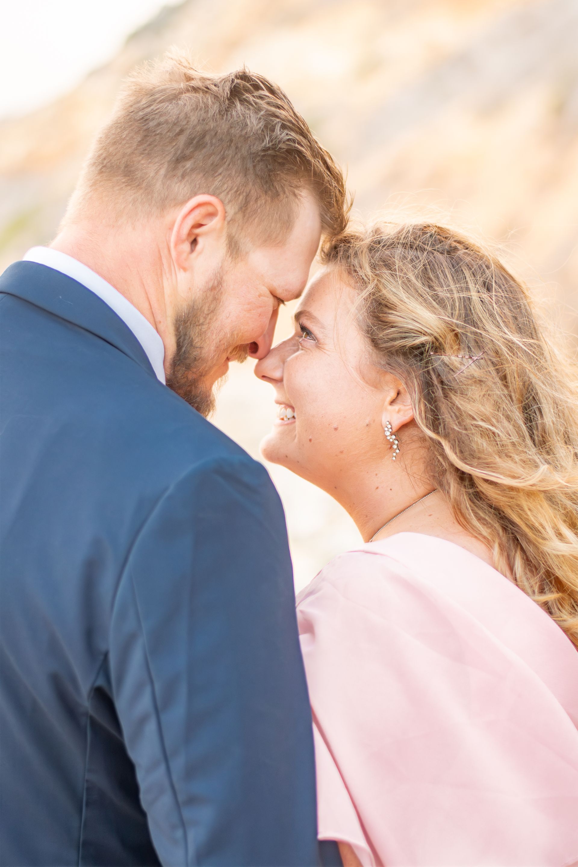 A couple in formal attire stands face-to-face, smiling as their foreheads touch outdoors.