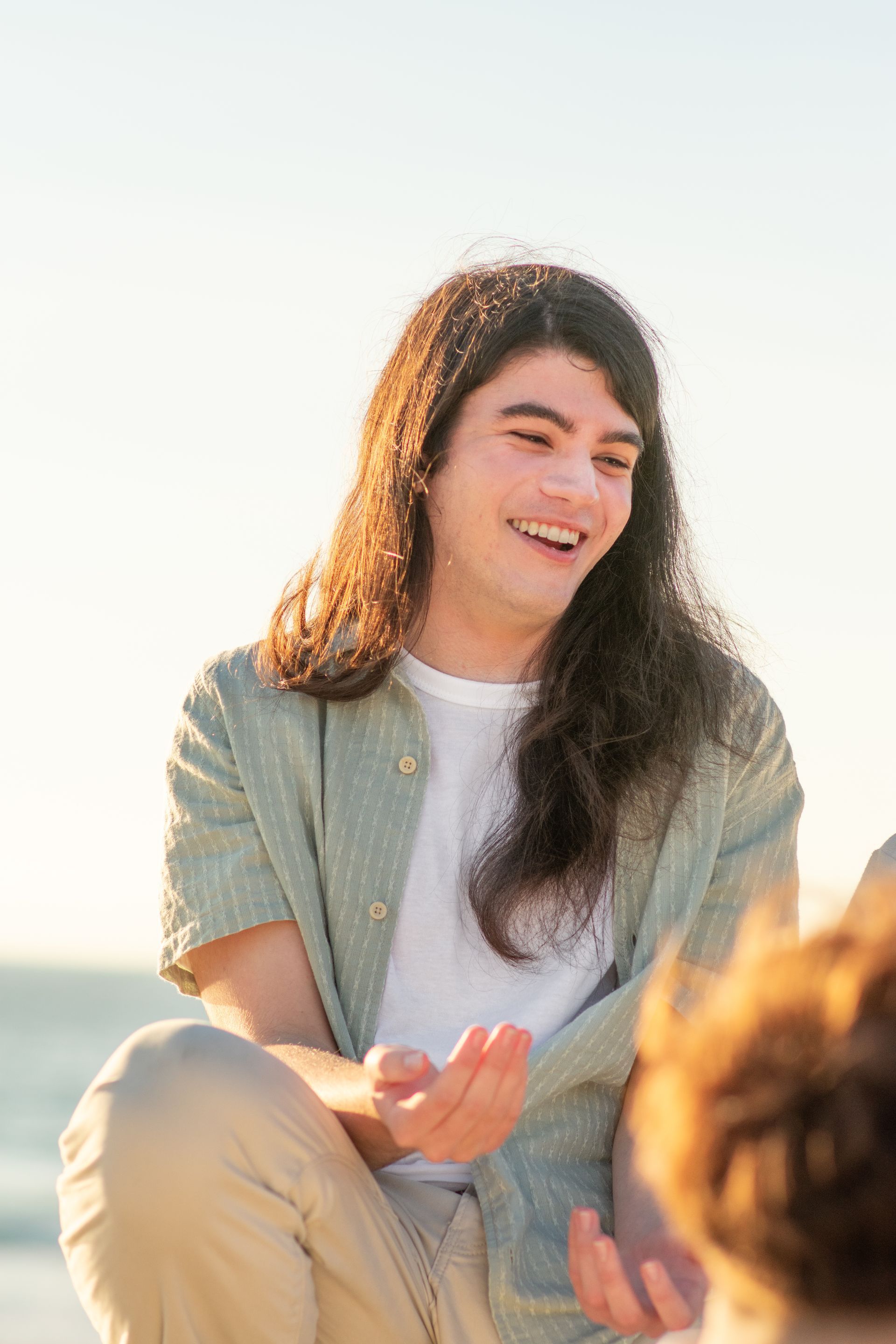 A young man with long hair is sitting on the beach talking to another man.