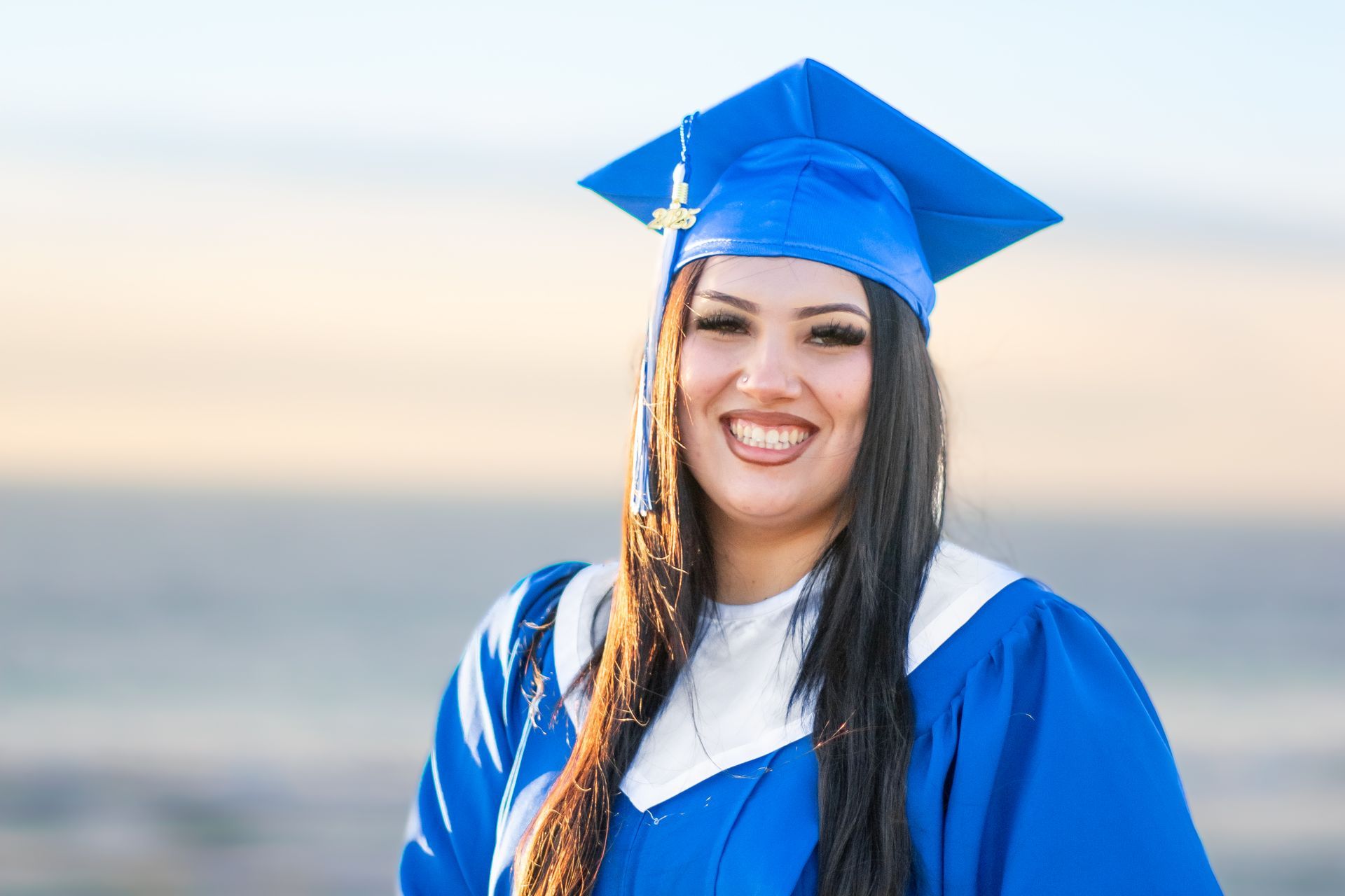 A woman in a blue graduation cap and gown is smiling at El Porto Beach.