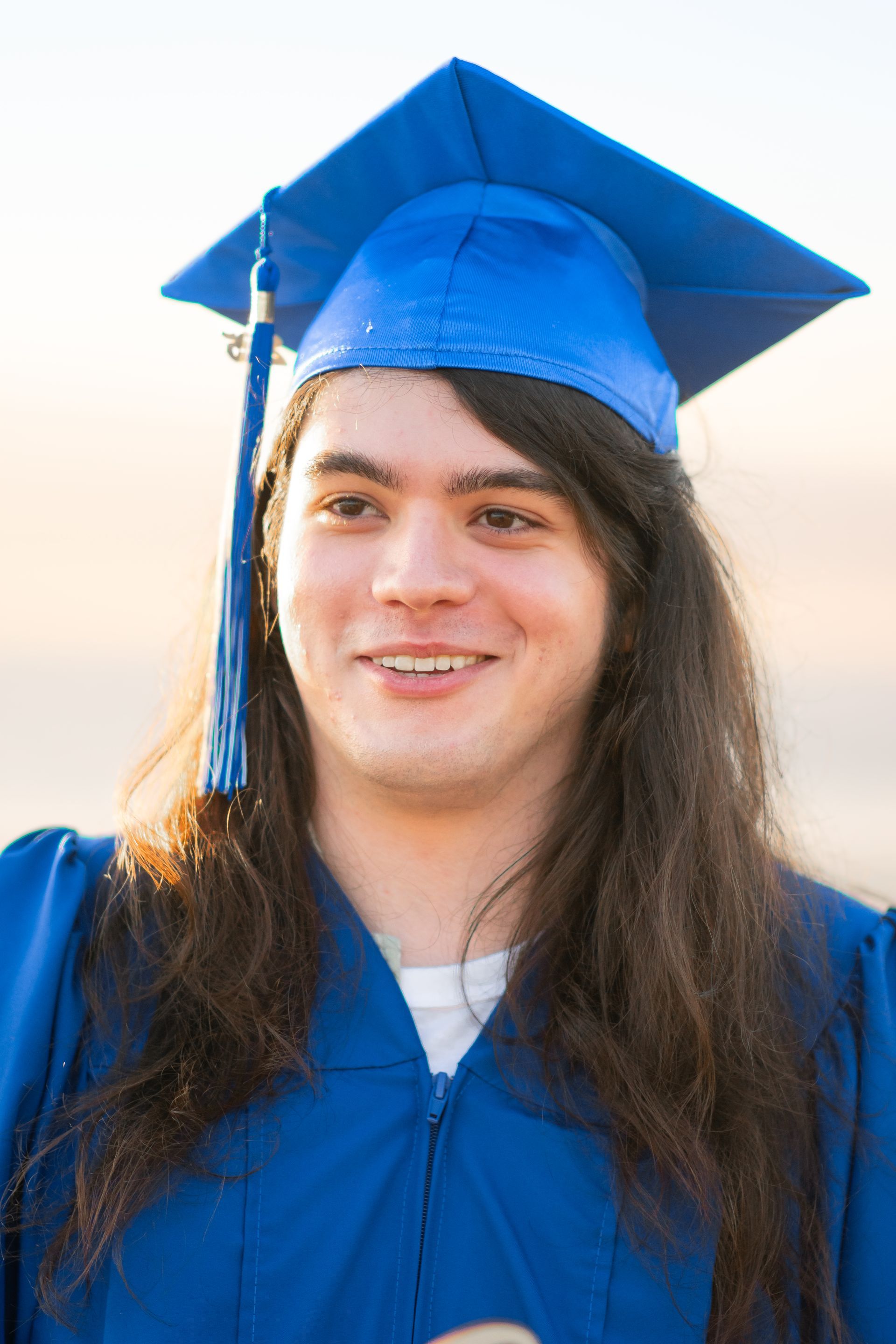 A young man with long hair is wearing a blue graduation cap and gown.