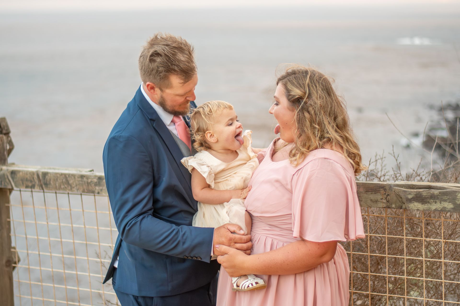 Family, mom and dad, with toddler making faces outdoors by a fence.