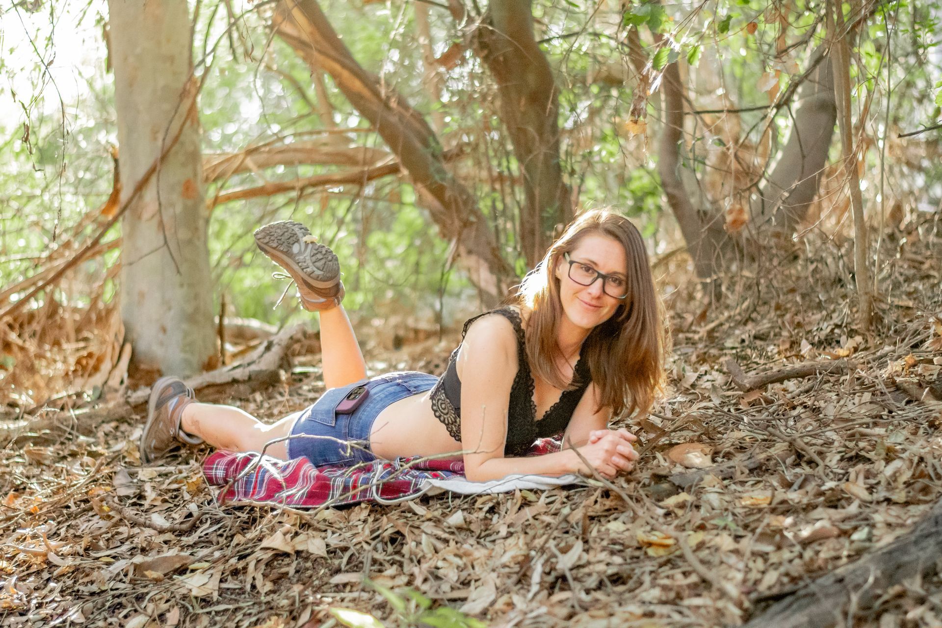 A woman is laying on her stomach in the woods reading a book.