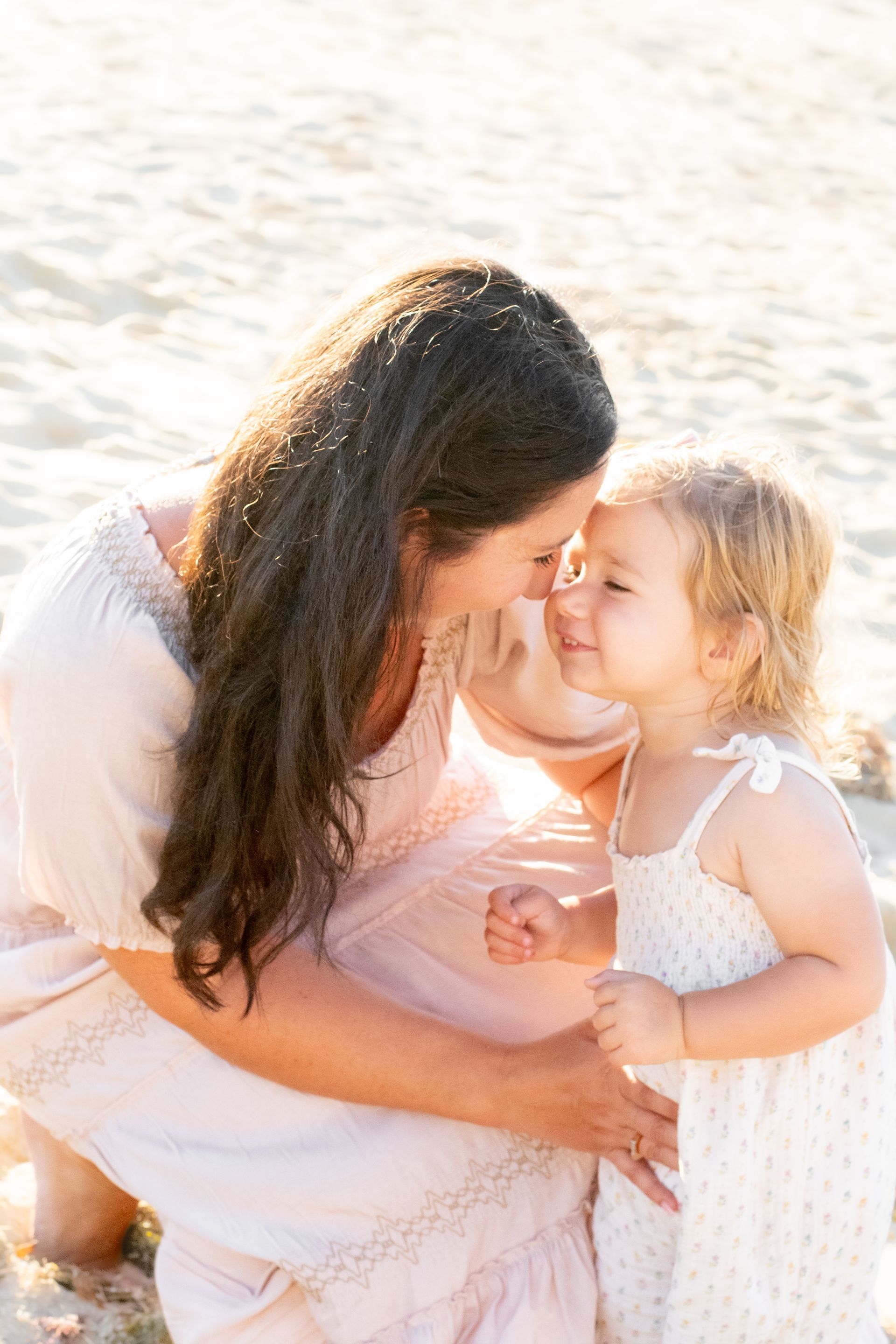 Mother and child touch noses on a beach, both smiling. Blonde toddler, dark-haired woman, sunny.