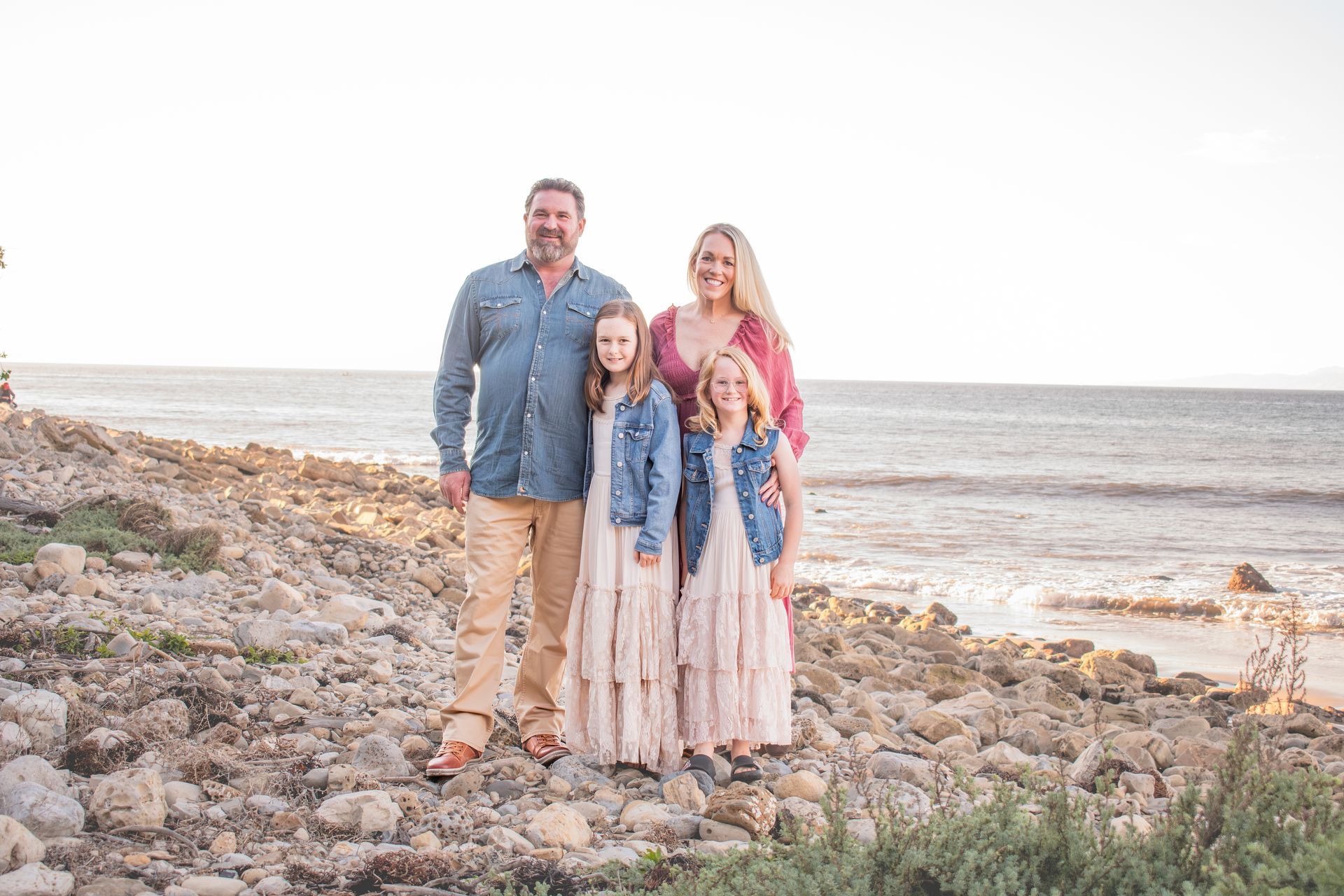 A family of four stands together on a rocky beach by the ocean during a sunny day.