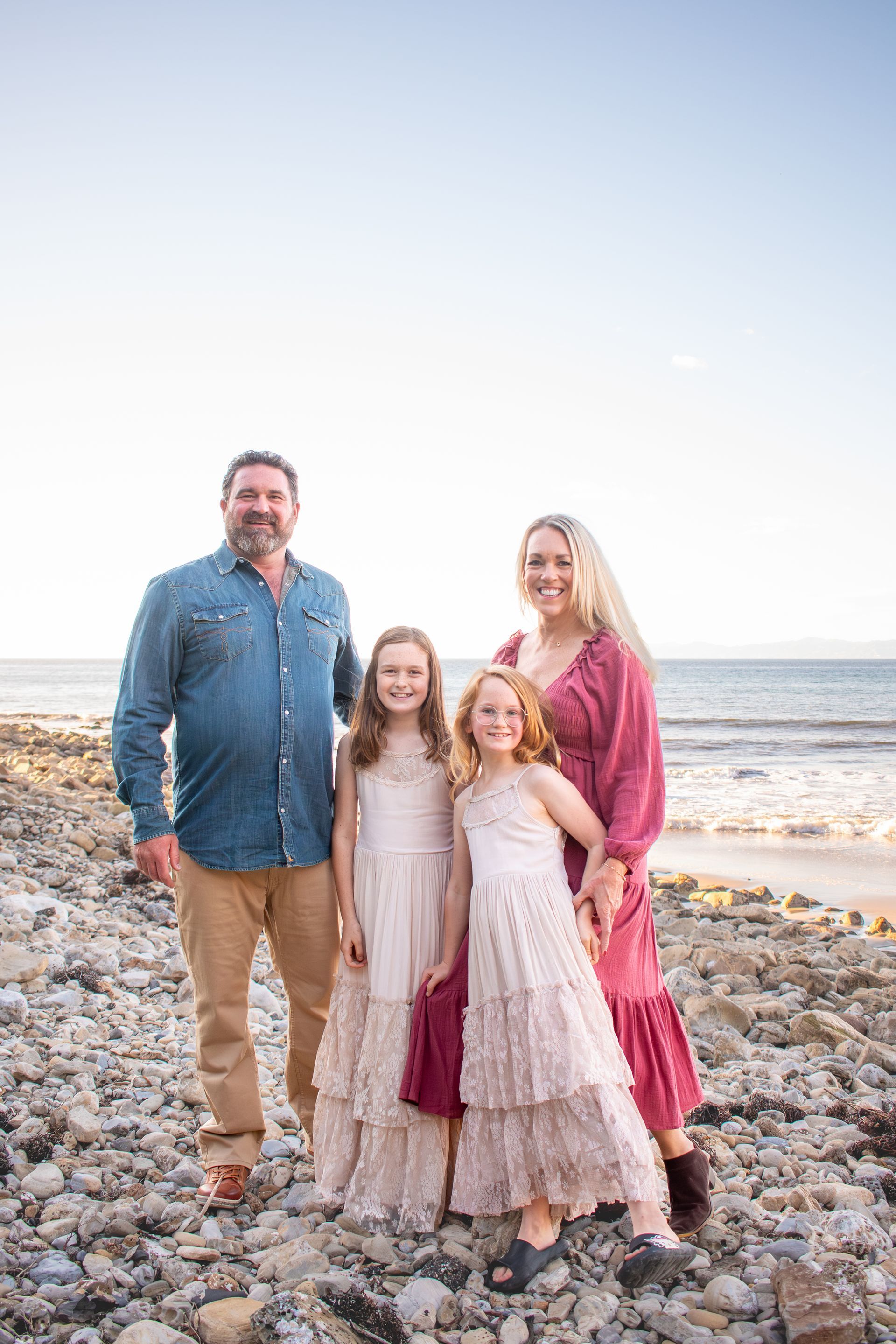 Family of four smiling on a rocky beach at sunset, wearing coordinated casual attire.