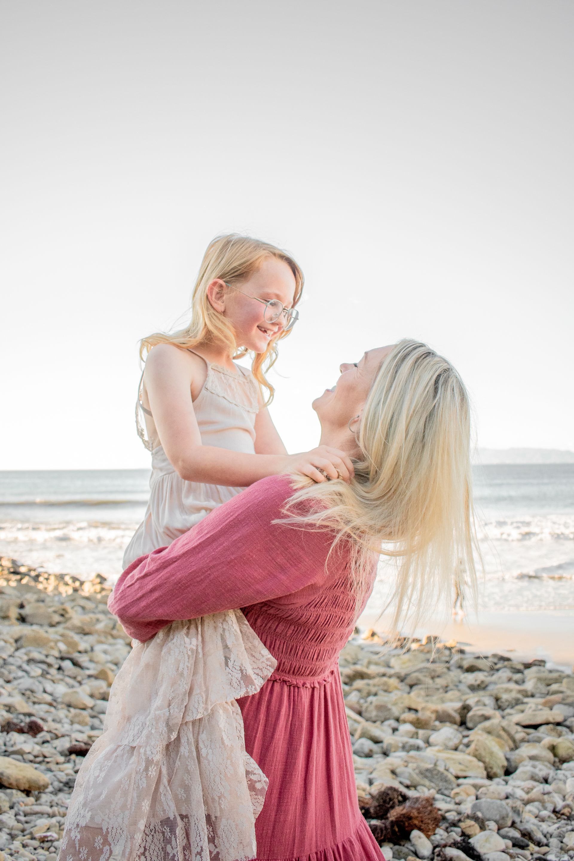 A person in a pink dress lifts a child in a cream-colored lace dress on a rocky beach, both smiling at each other.