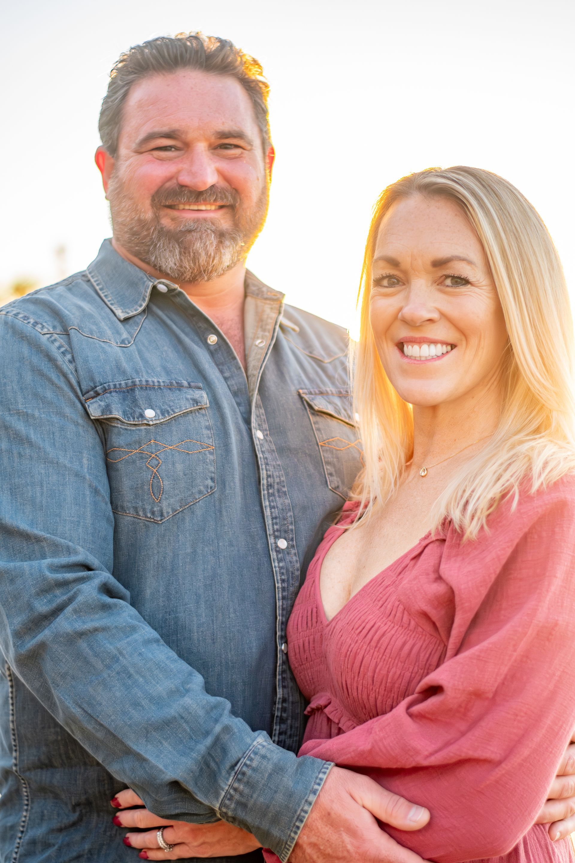 A smiling couple embrace during a sunny outdoor portrait session; he wears a denim shirt, she wears a reddish-pink top.