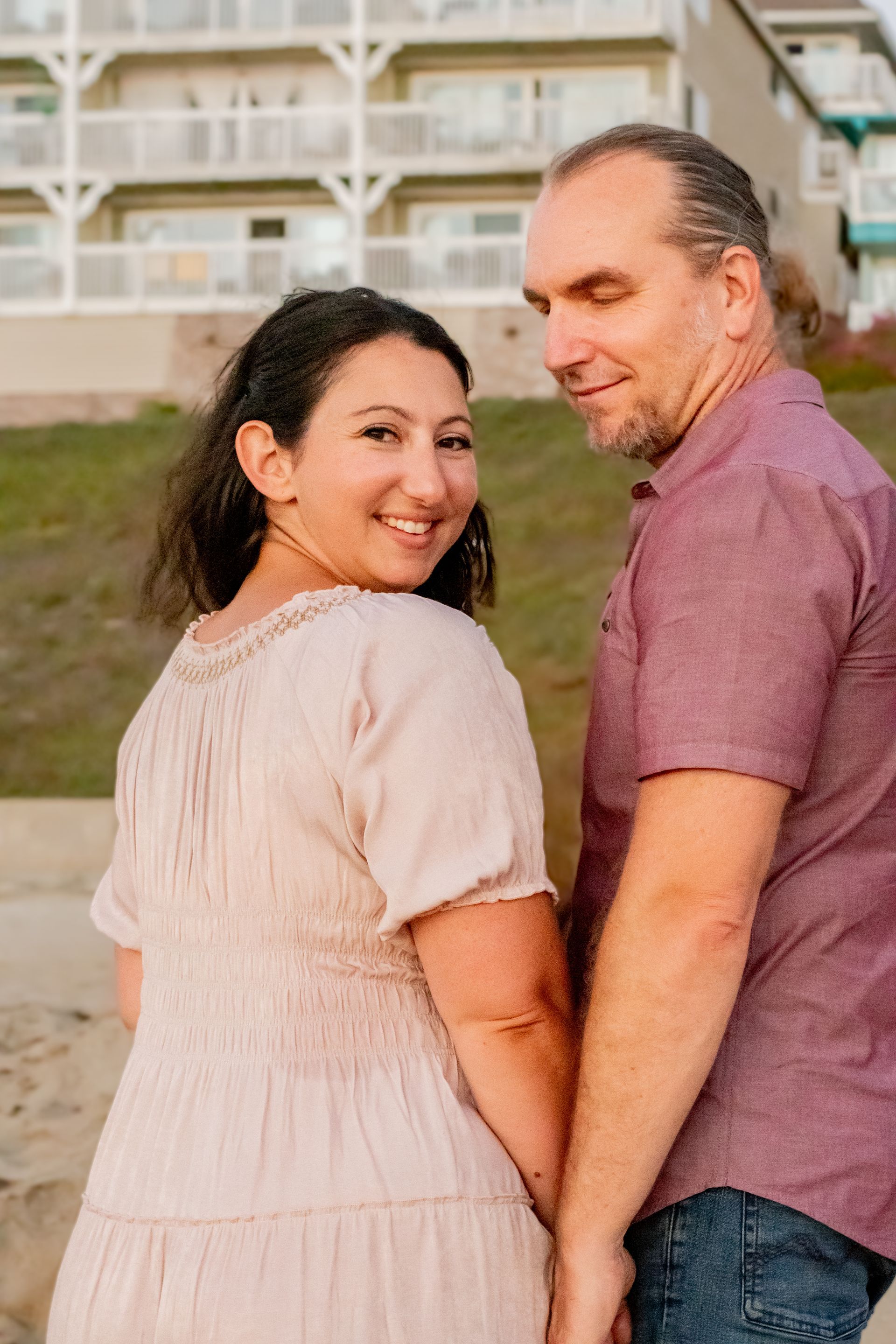 Woman and man holding hands, smiling at the camera on a beach, with a building in the background.