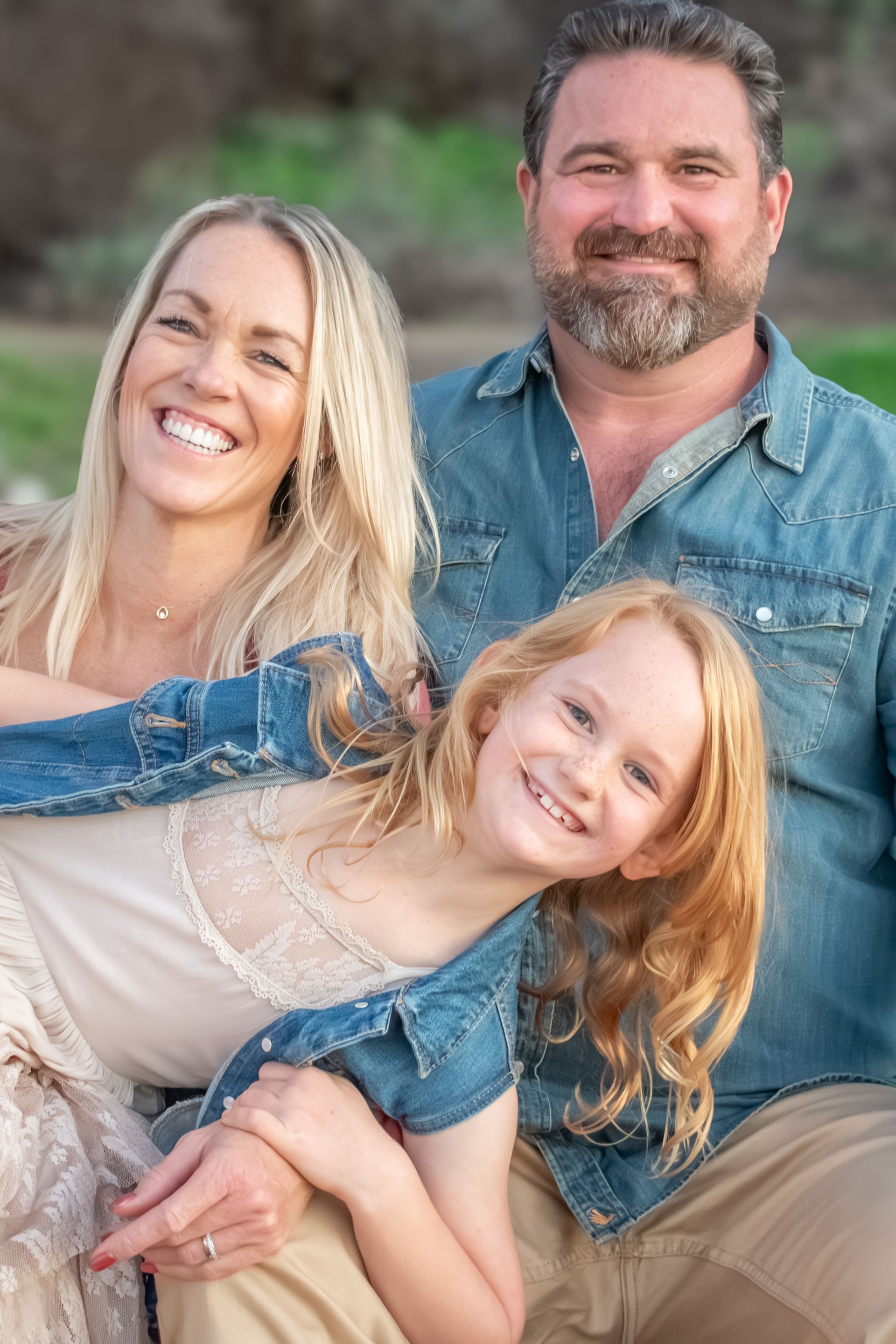 A smiling family of three posing outdoors for a portrait, with a man, woman, and child huddled close together.