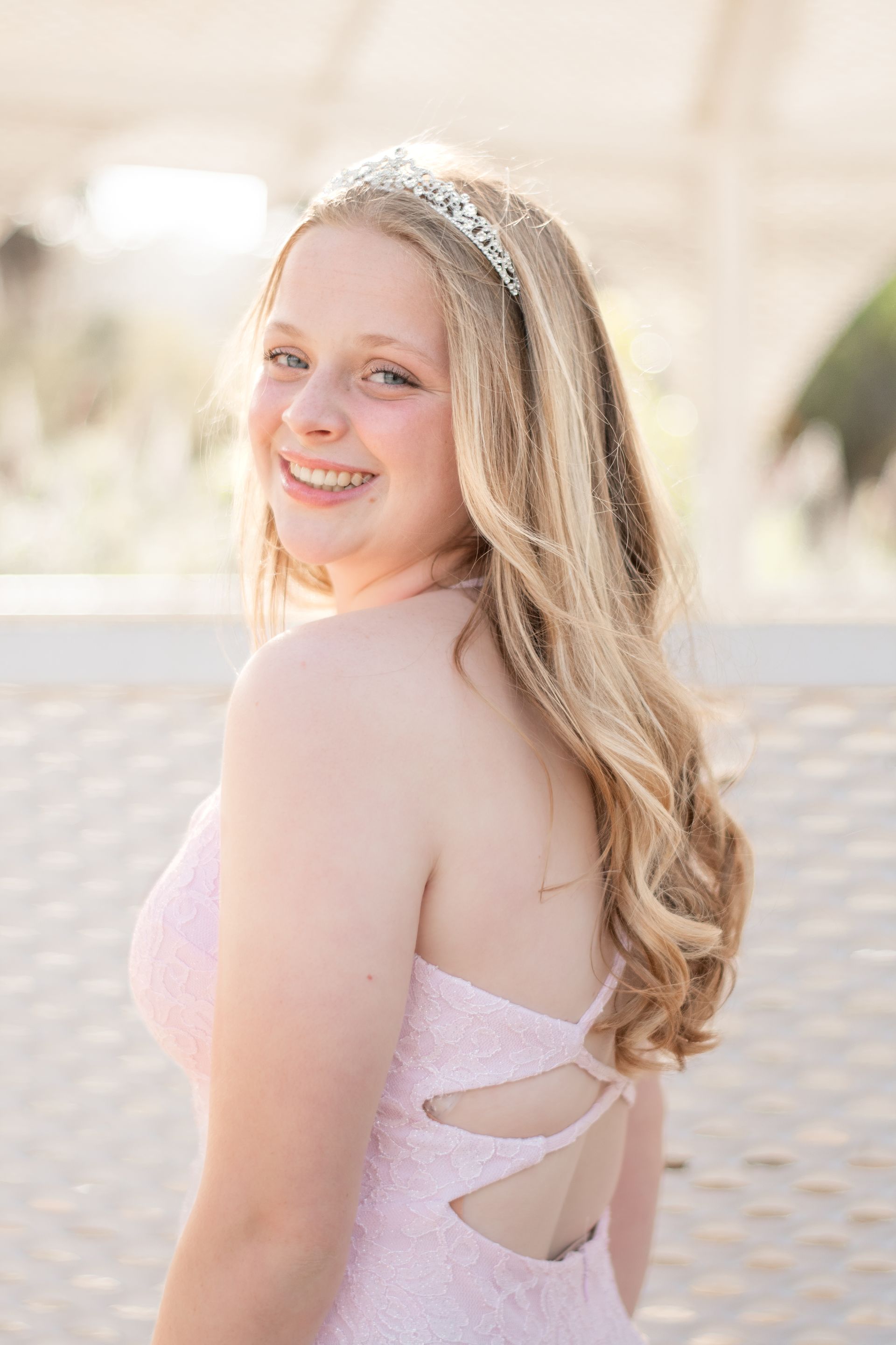 Blonde girl in a pink dress and tiara smiles over her shoulder at an outdoor event.