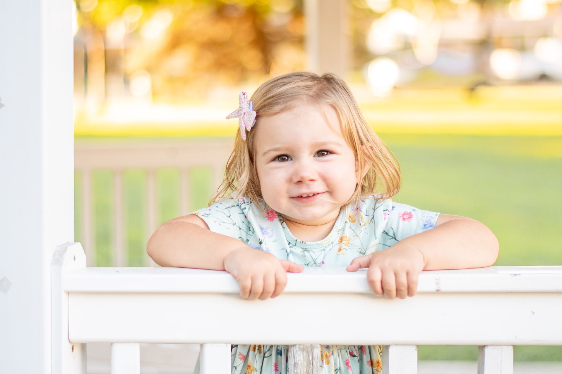 A young child with a hair clip leans over a white railing in a grassy, sunlit outdoor setting.