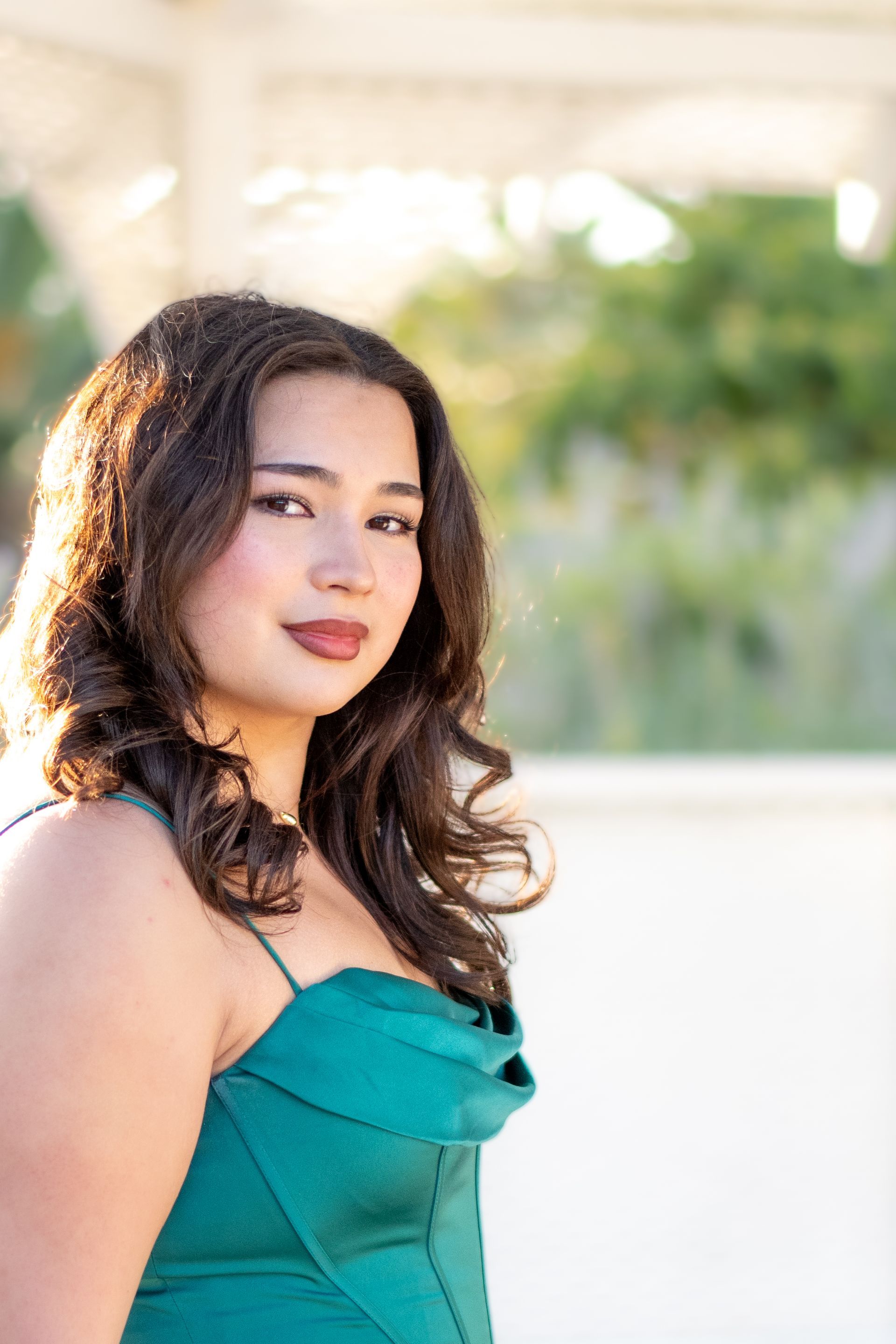 Woman in teal dress smiles, with long dark hair, outdoors in front of a gazebo.