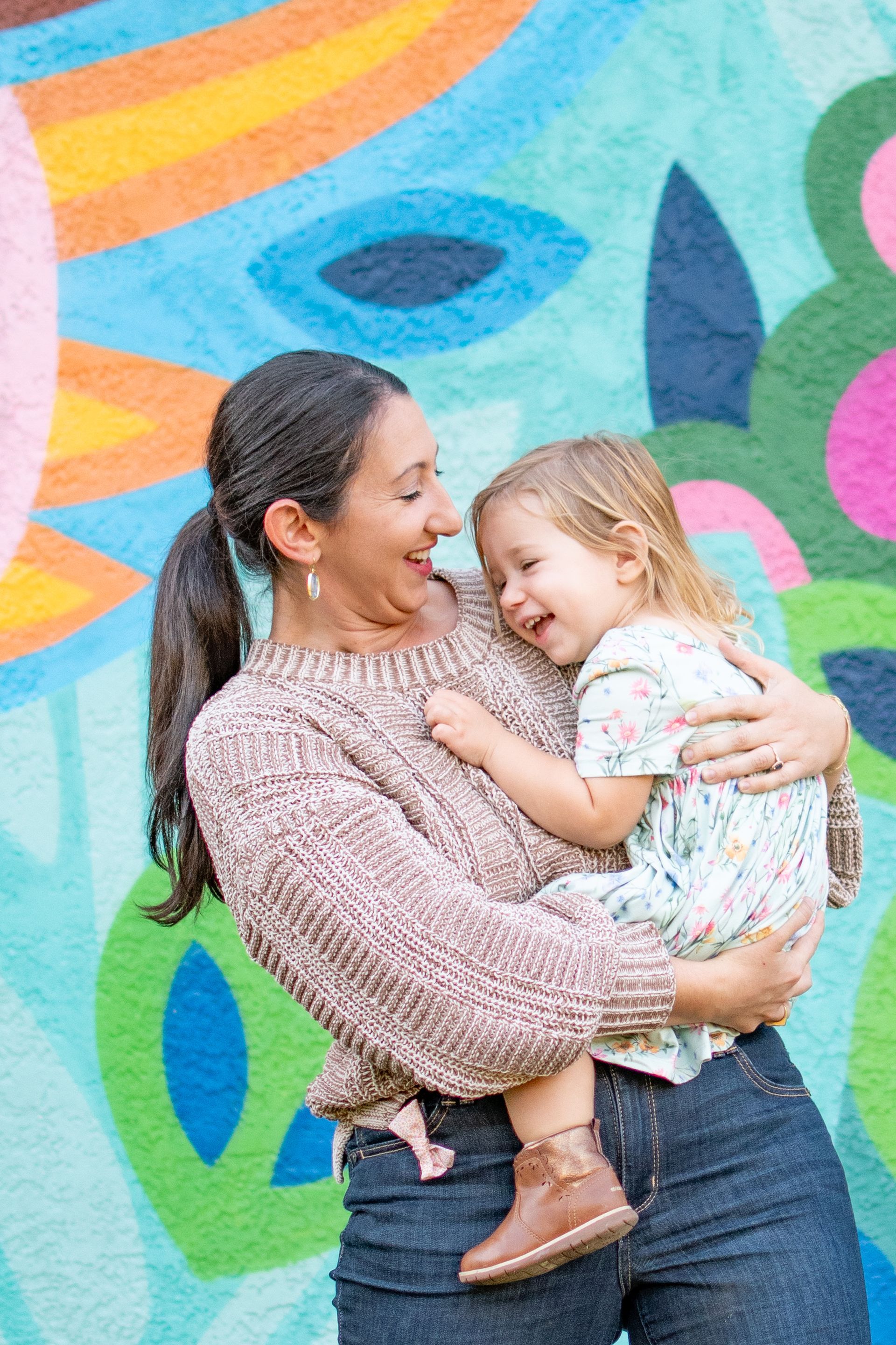 A person in a brown sweater holds a smiling child in a floral dress in front of a colorful, abstract mural.