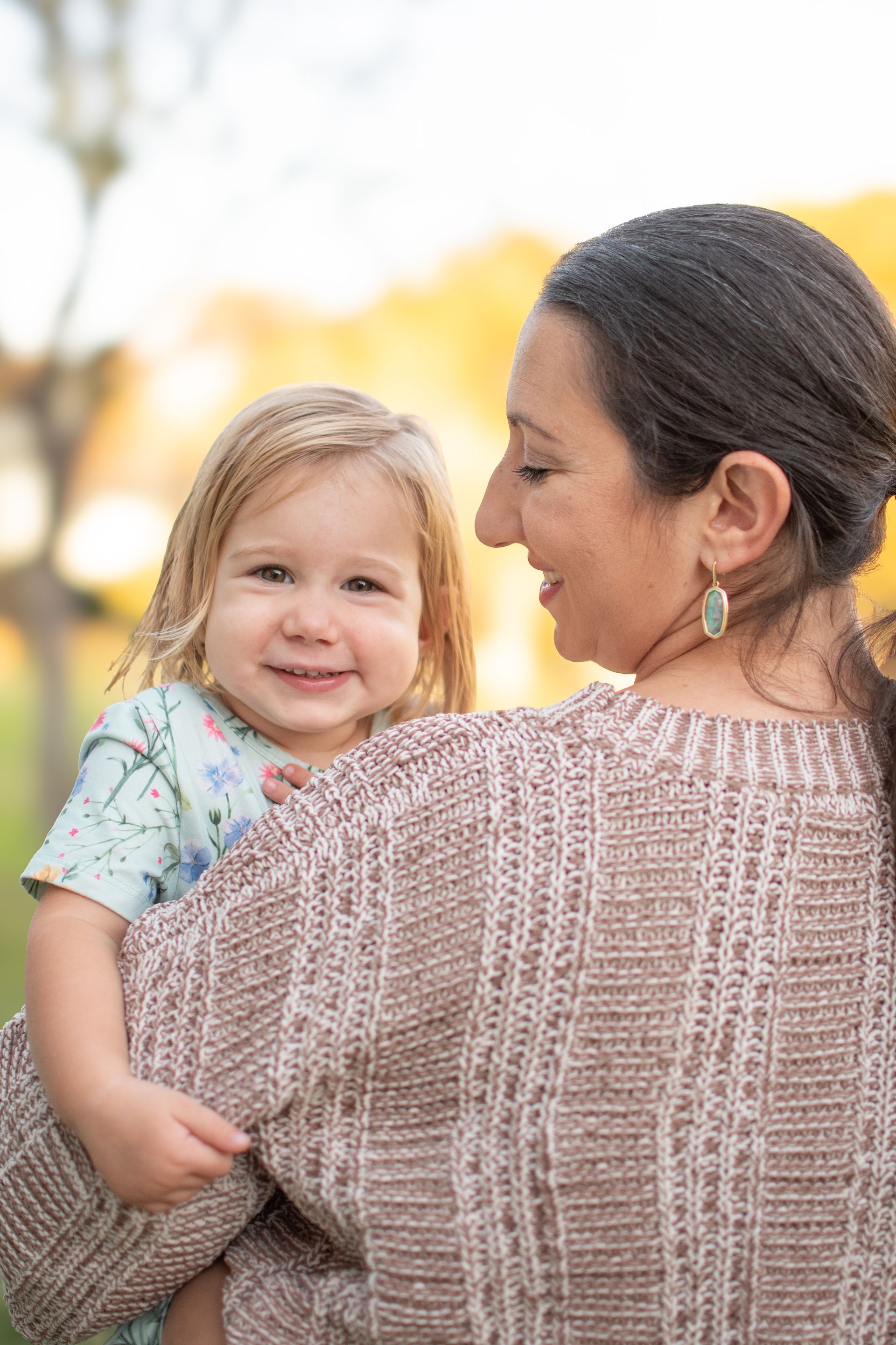 A person in a textured tan sweater holds a smiling toddler in a patterned light blue shirt against a blurred outdoor backdrop.