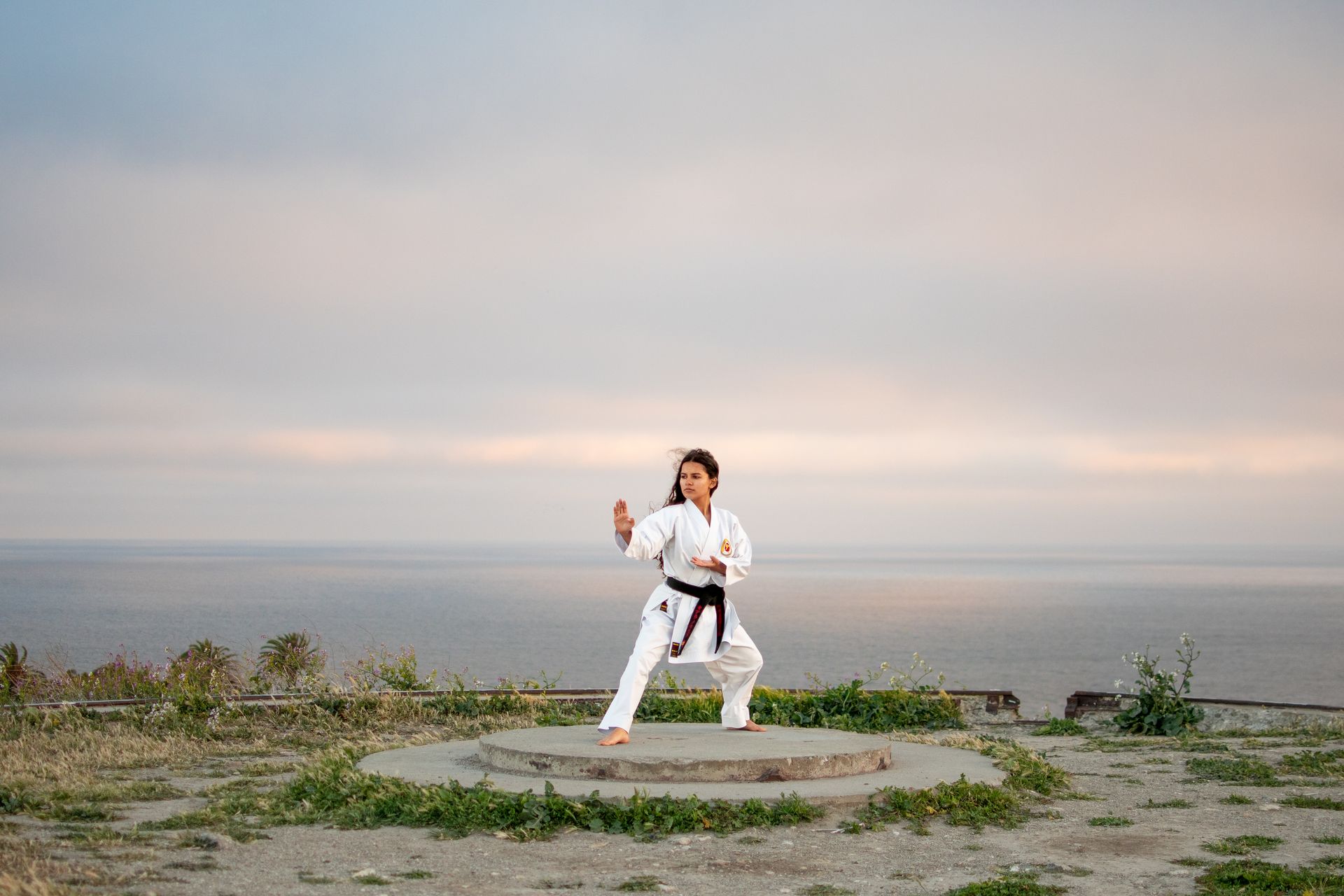 A person in a white karate gi and black belt performs a martial arts stance on a circular stone platform overlooking a sea.
