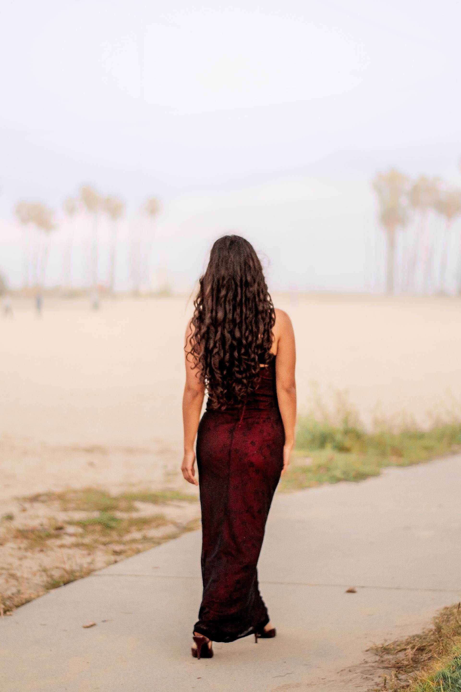 A woman in a long red dress is walking down a sidewalk on a South Bay Beach