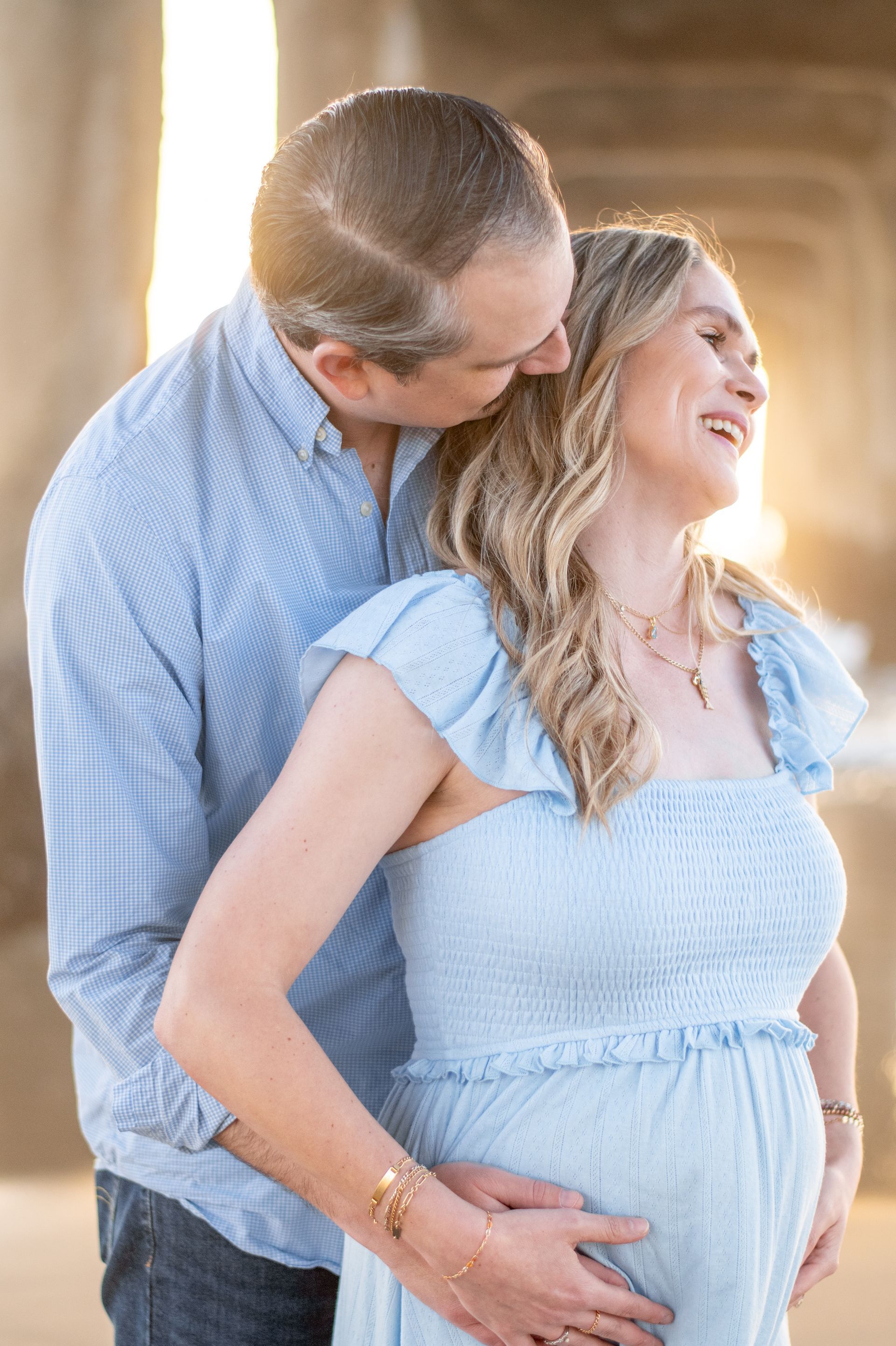 A person in a blue dress and a person in a blue shirt embrace outdoors, backlit by warm, golden sunlight.