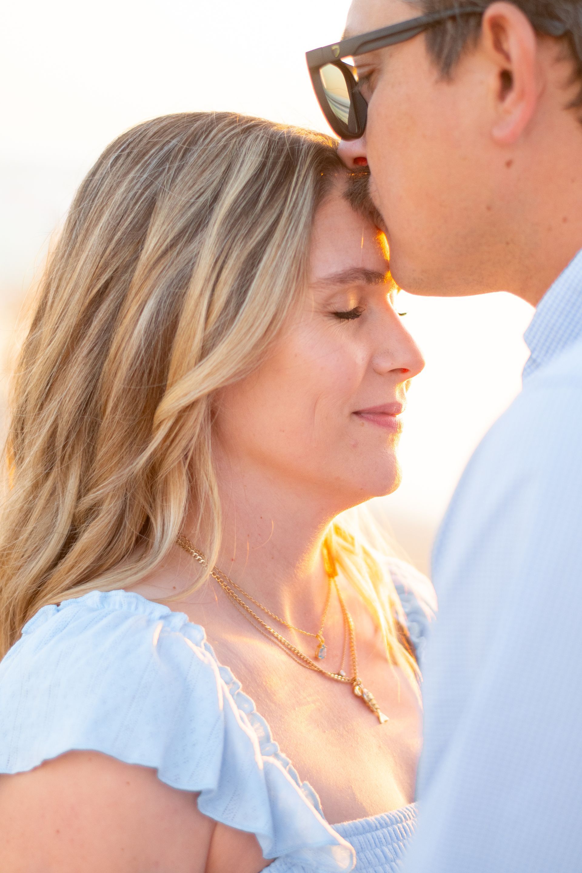 A person in a light blue top with closed eyes receives a gentle kiss on their forehead from a partner wearing sunglasses.
