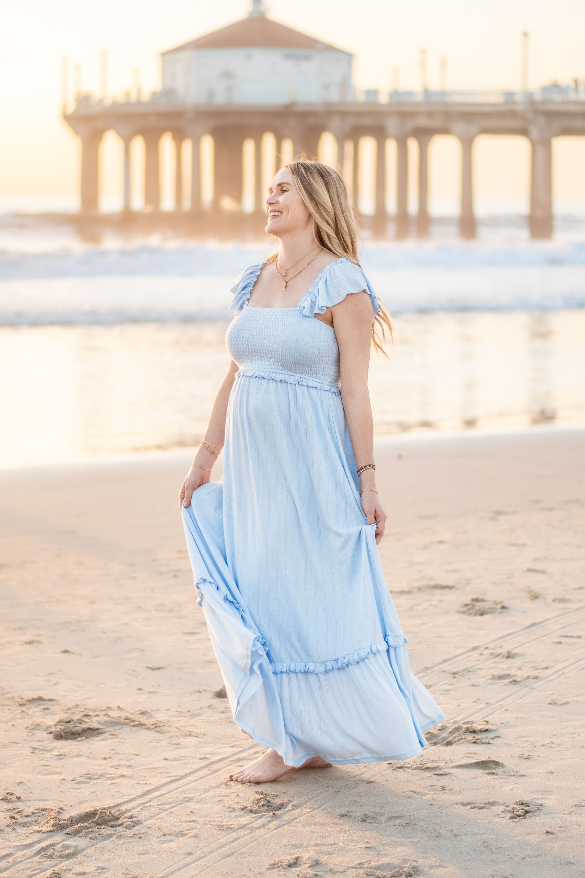 A person in a light blue sundress walking barefoot on a beach with a pier in the background during golden hour.