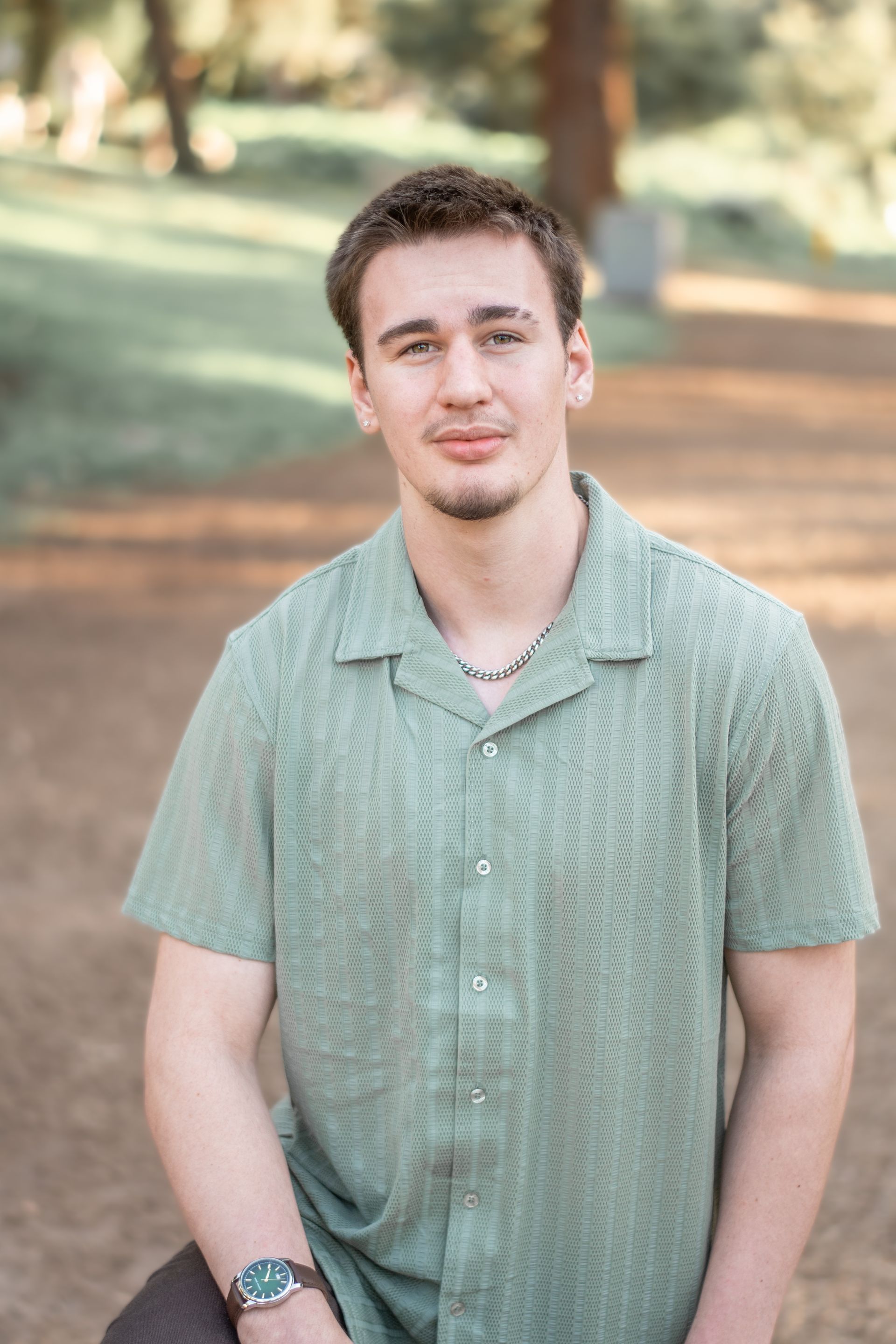 A person wearing a textured green shirt and a necklace poses outdoors in a sunlit, wooded setting.