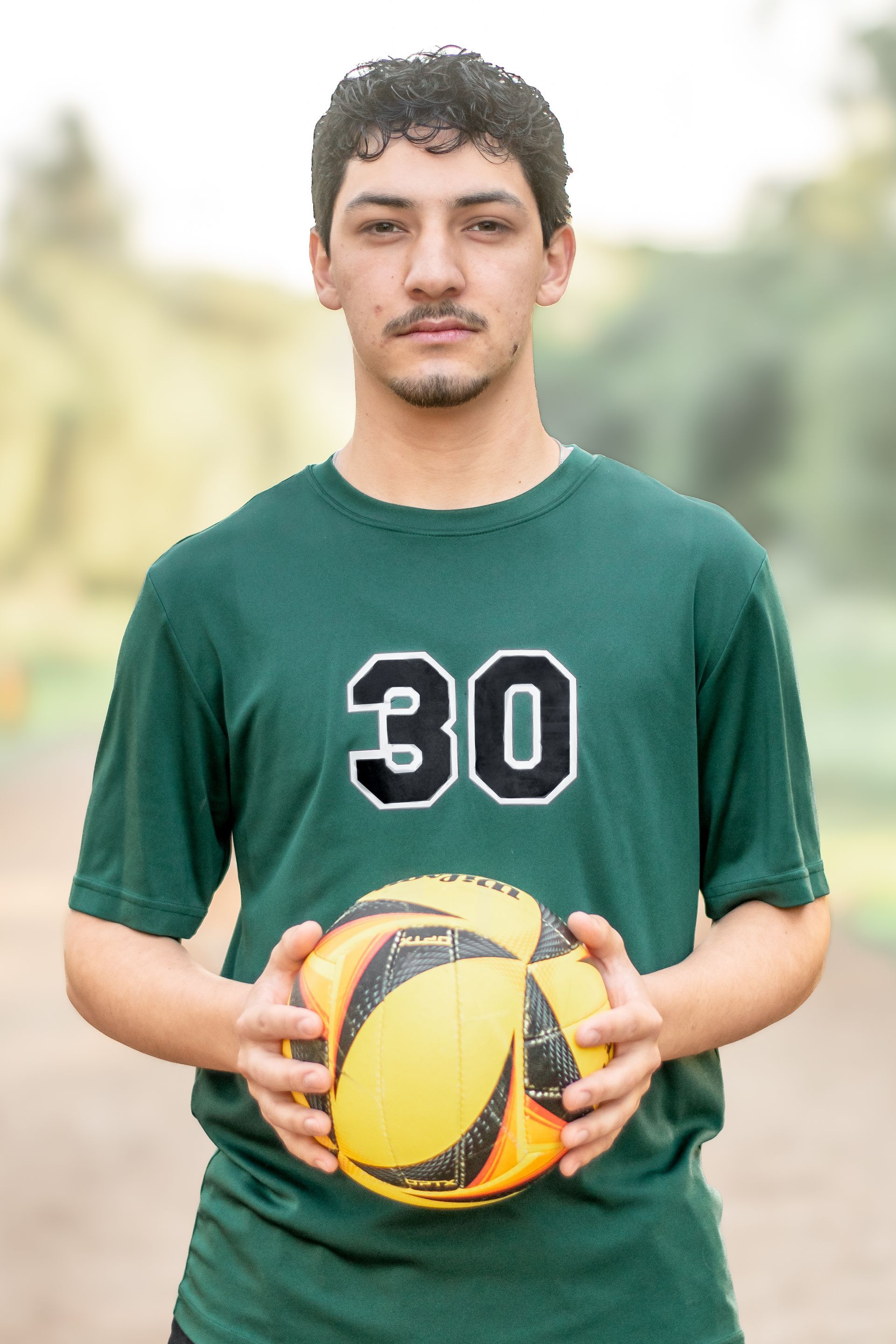 A person with dark hair wearing a green shirt with the number 30, holding a yellow and black ball outdoors.