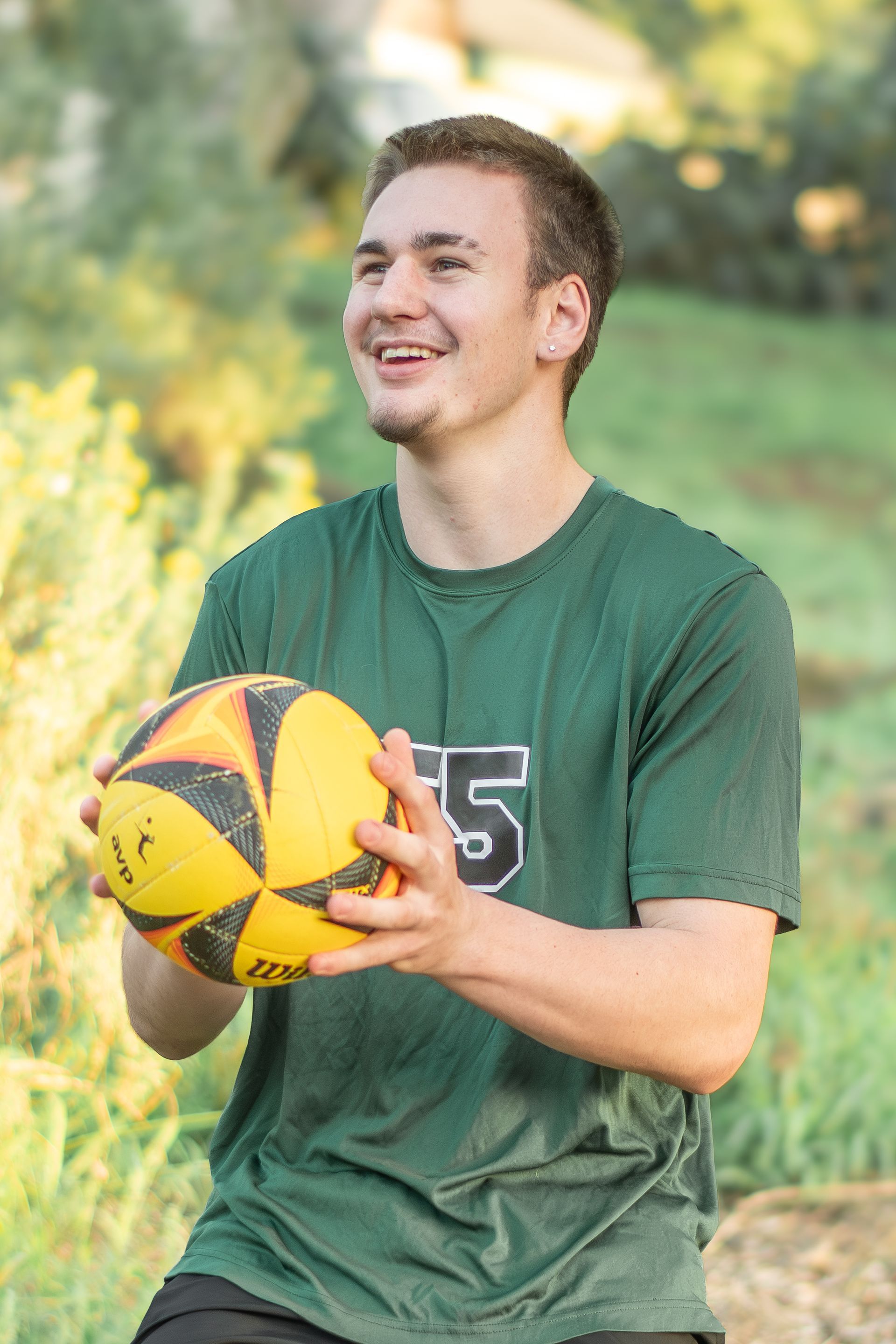A smiling person in a green shirt holds a yellow and black volleyball outdoors with greenery in the background.