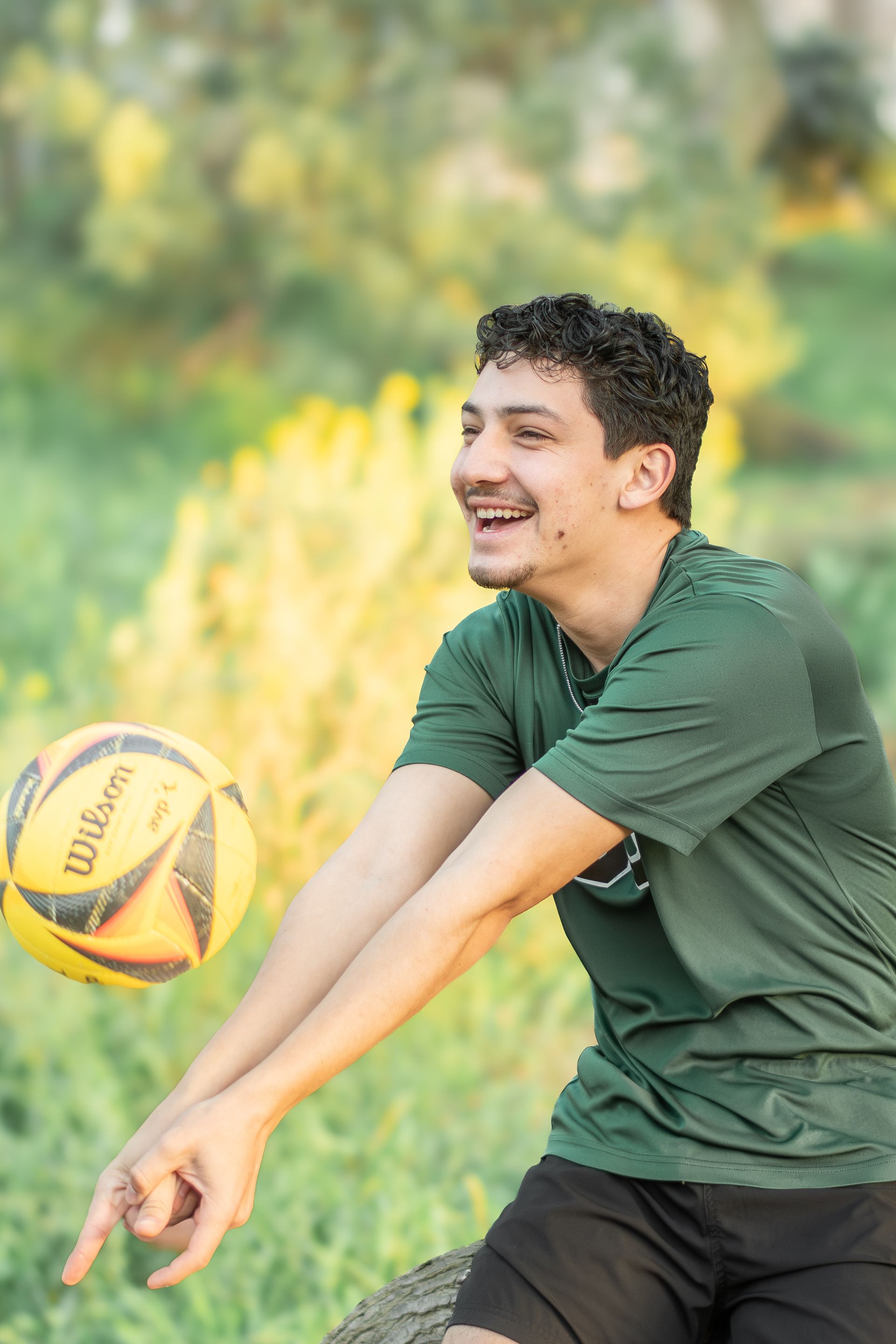 A smiling person in a green shirt practicing a volleyball forearm pass outdoors in a grassy area.