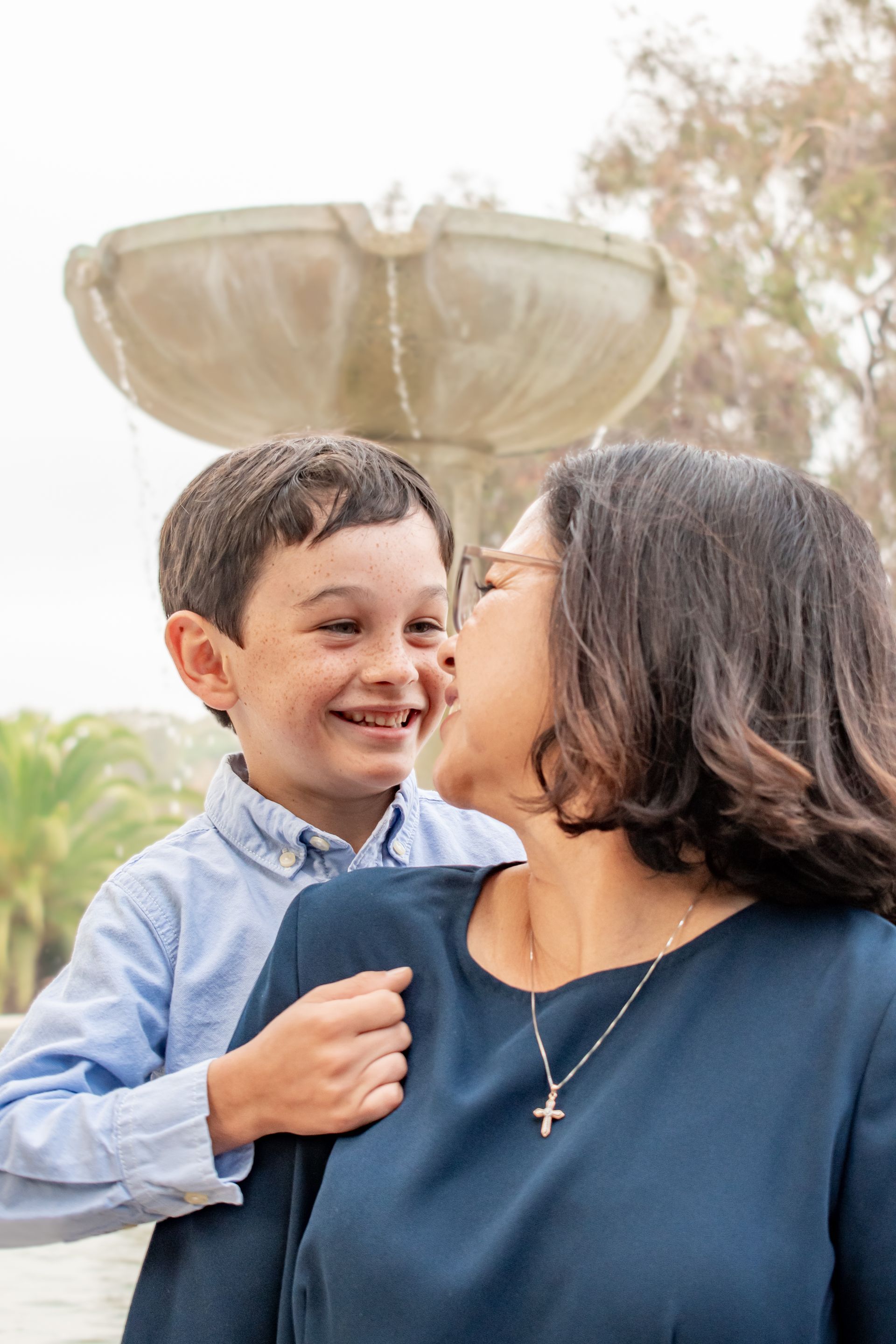 A smiling child leans toward a woman wearing a blue top and a cross necklace, with a stone fountain in the background.