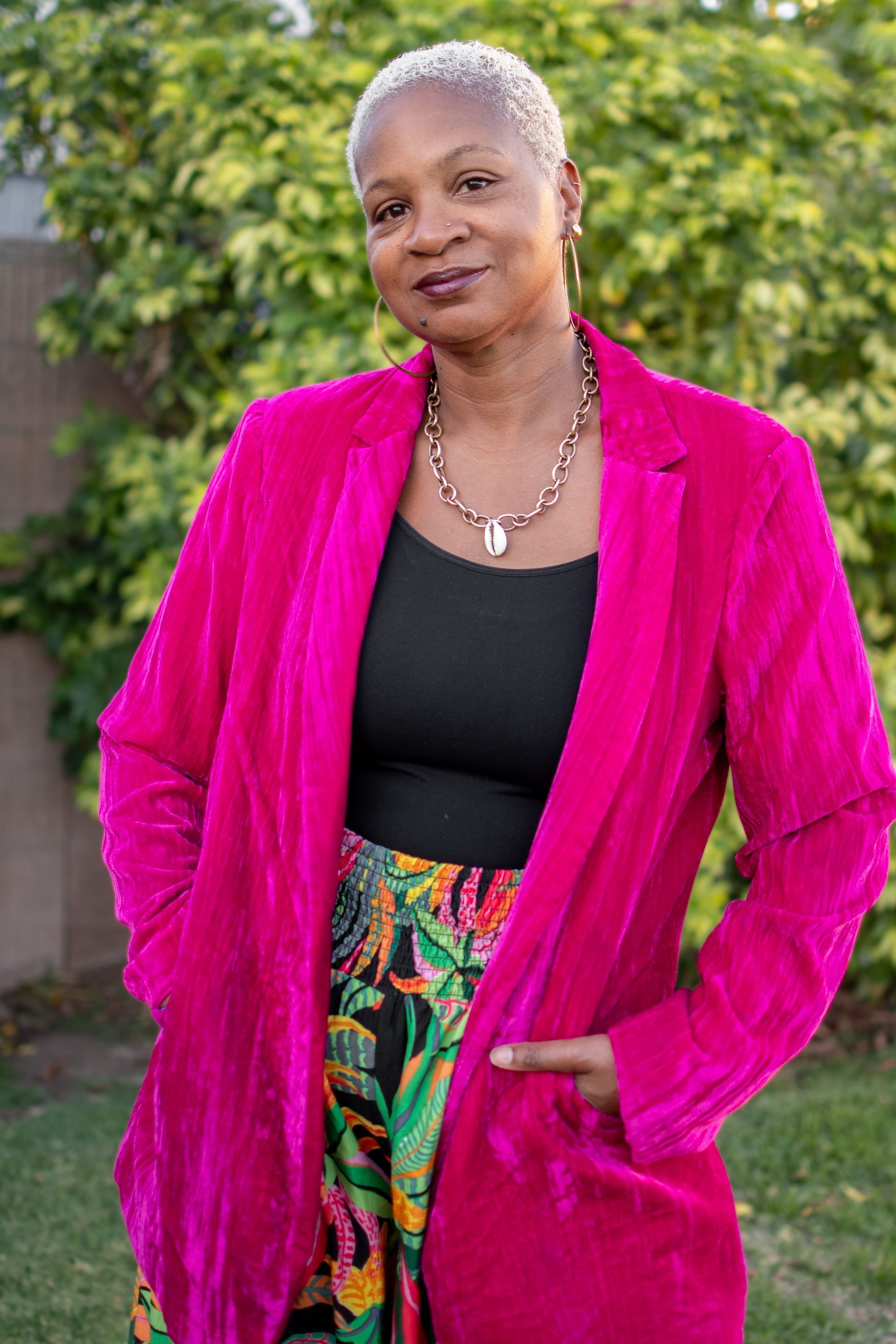 A person with short, light-colored hair wearing a vibrant pink blazer, black top, and patterned skirt in a garden.
