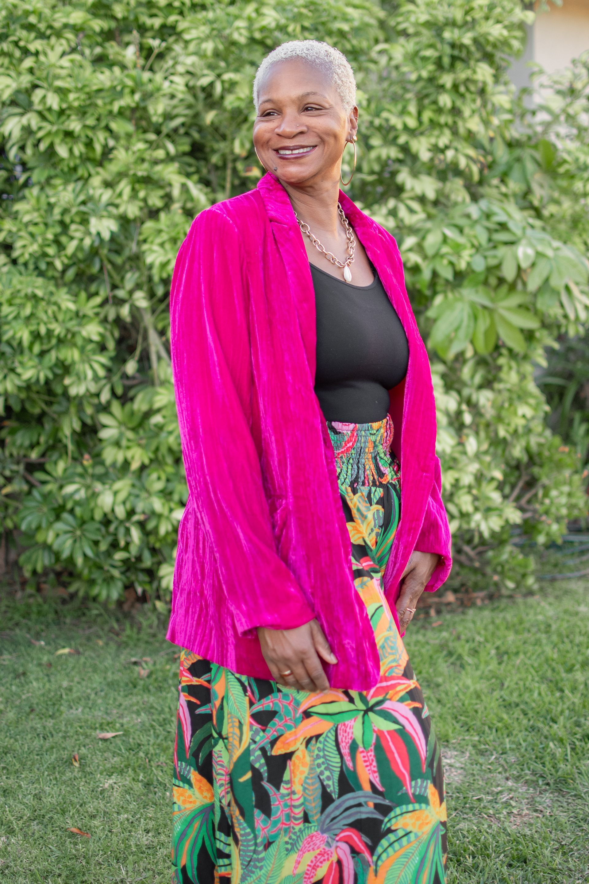 A person with short, light-colored hair wearing a bright pink velvet blazer and a tropical print maxi skirt outdoors.