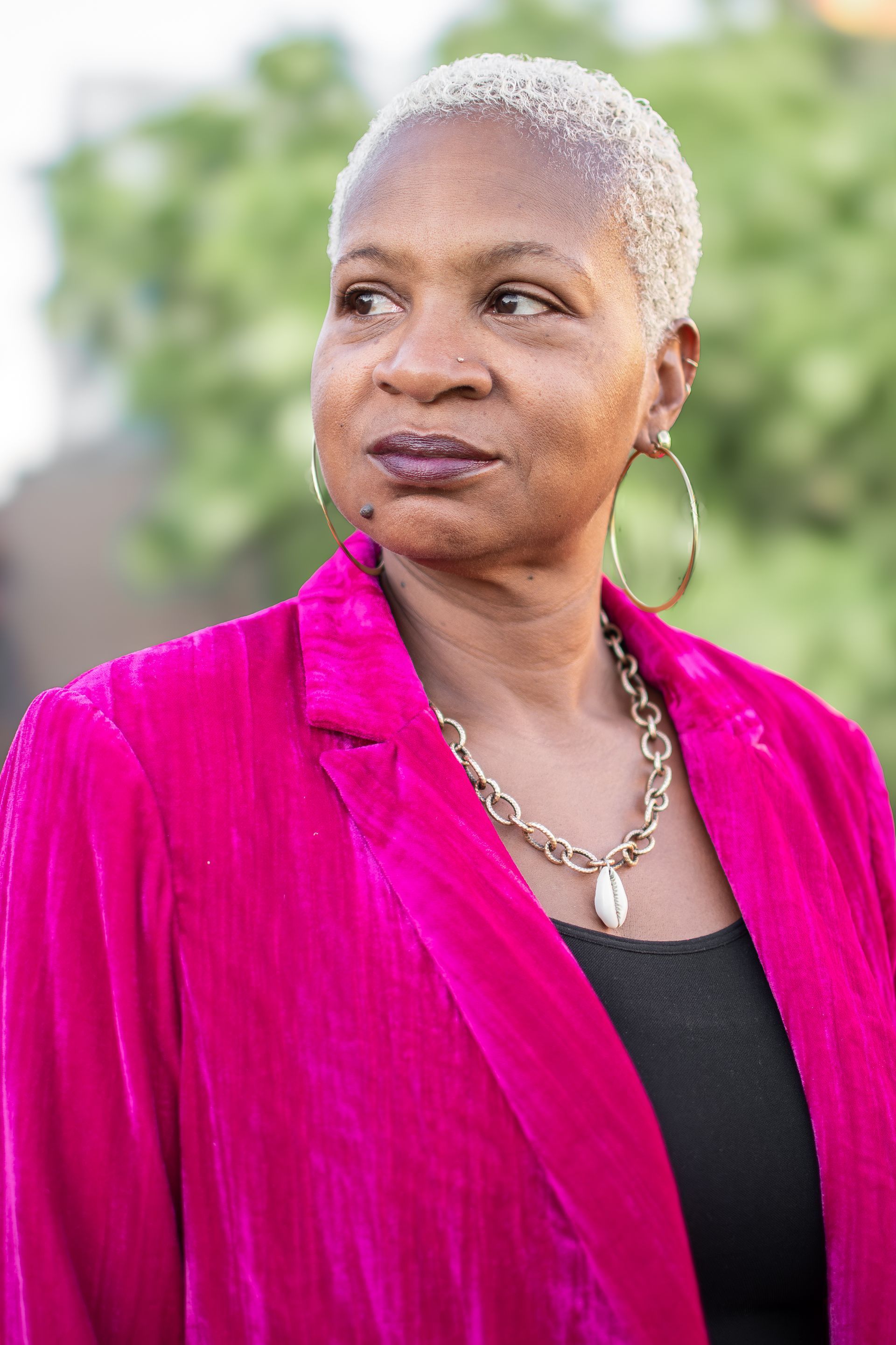 A person with a cropped light hairstyle wears a bright pink blazer, large hoop earrings, and a gold chain necklace.