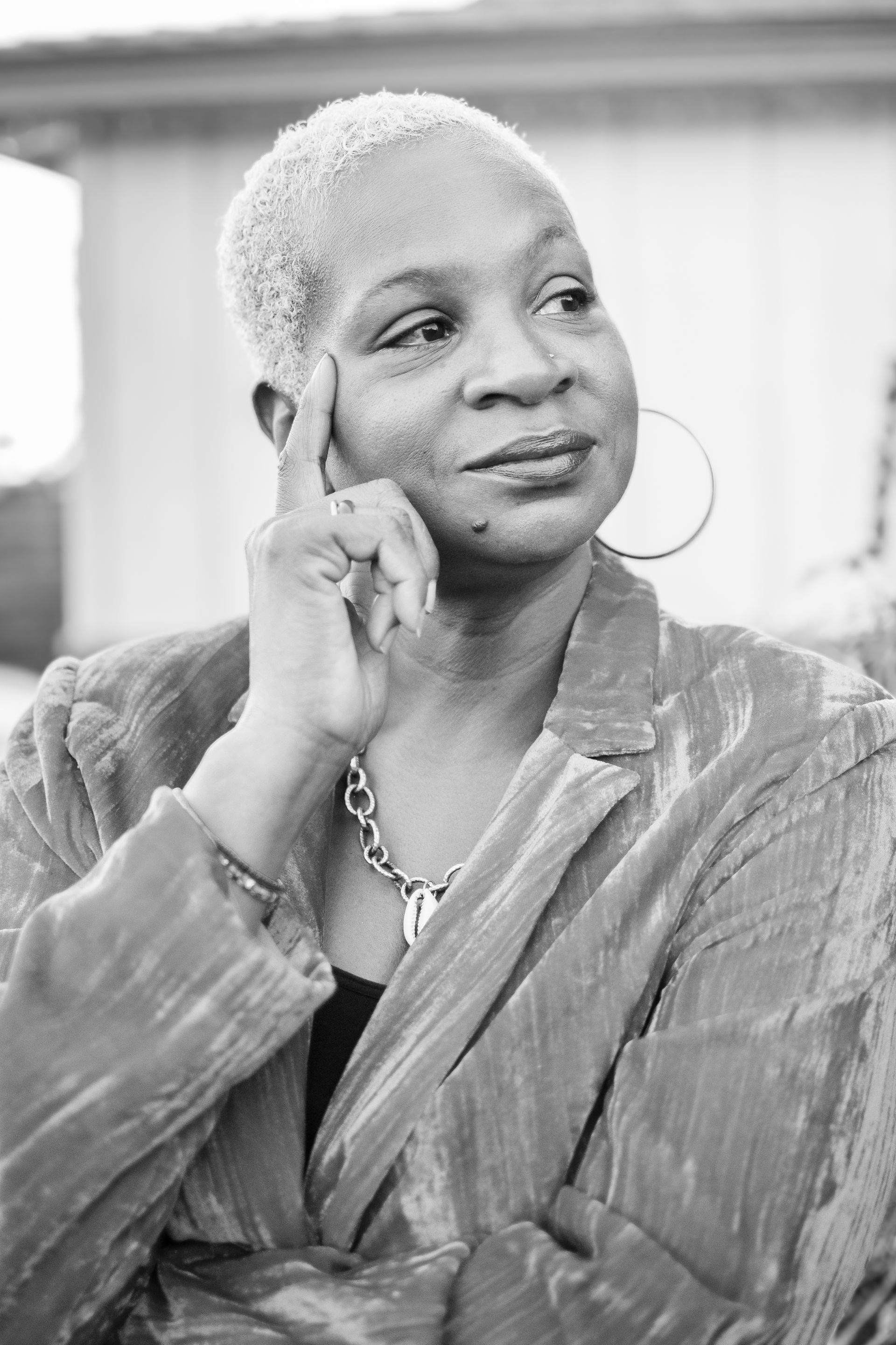 A person in a textured blazer with a necklace, resting their temple on a finger while looking off-camera.