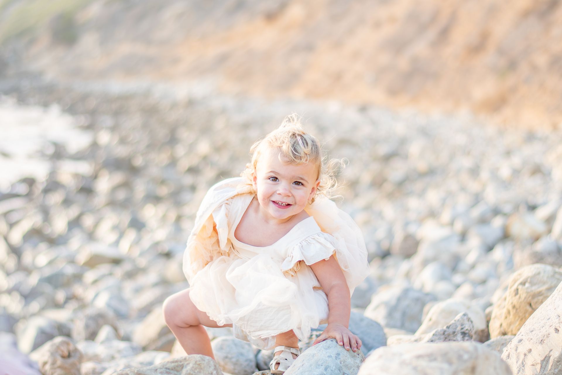 A young child in a cream dress climbing over smooth rocks on a beach at sunset.