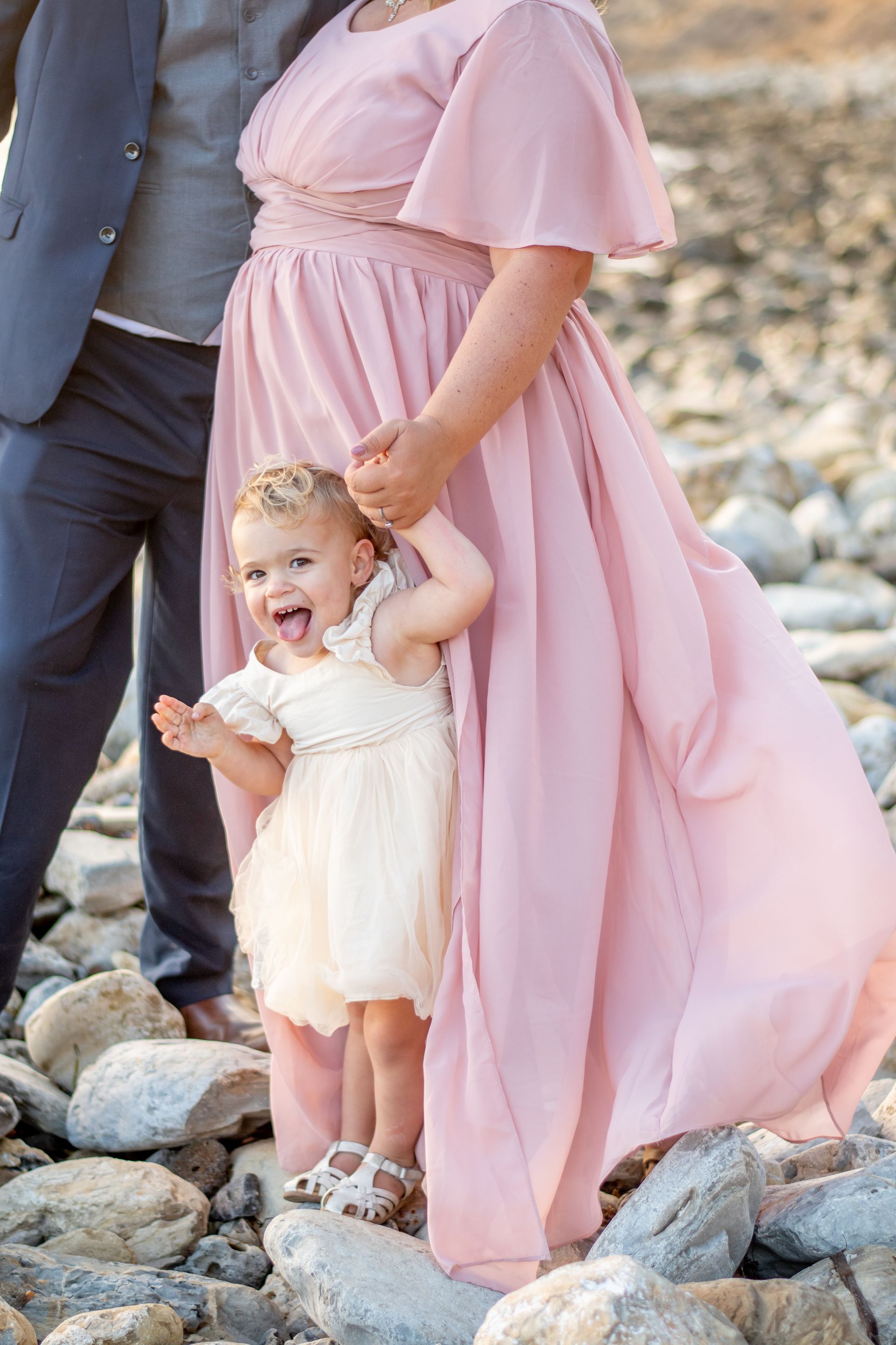 A toddler stands on rocks while holding the hand of a person in a long, flowing pink dress, with another person in a suit.