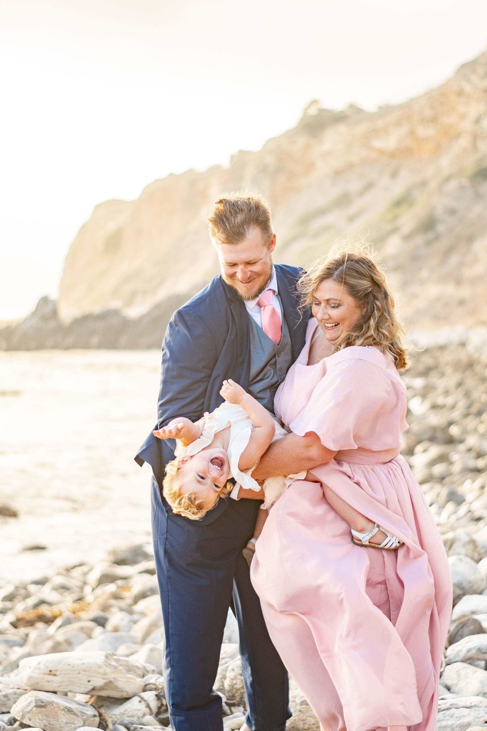 A person in a blue suit and a person in a pink dress hold a small child upside down on a rocky beach at sunset.
